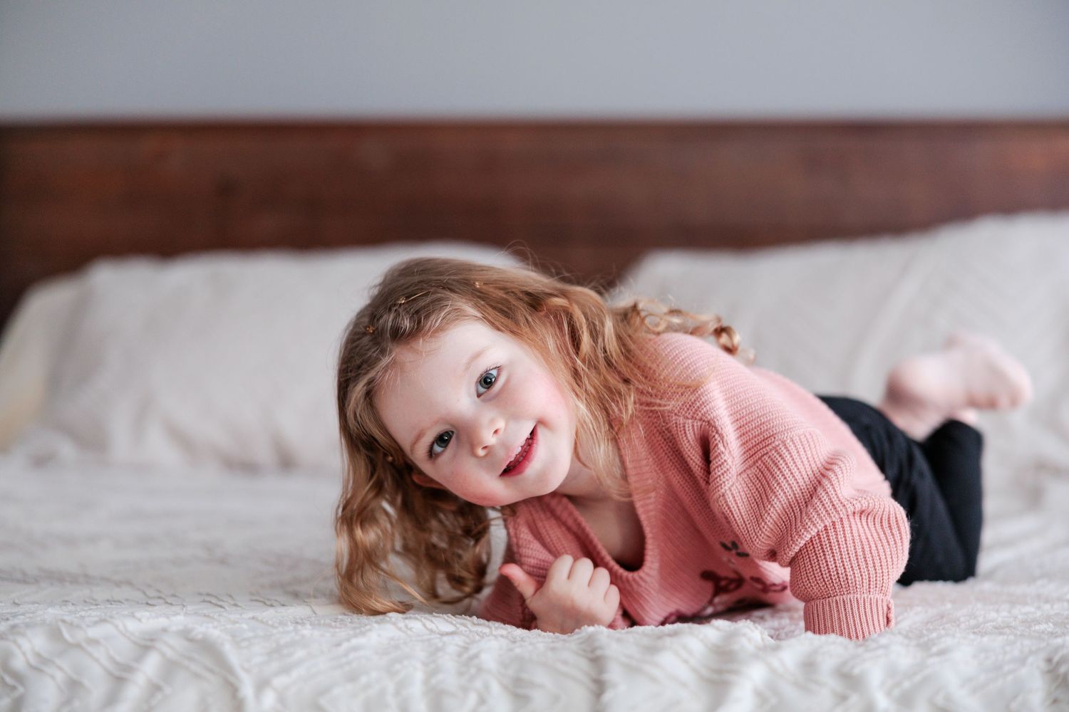 Adorable toddler girl with curly hair smiling while lying on a white bed.