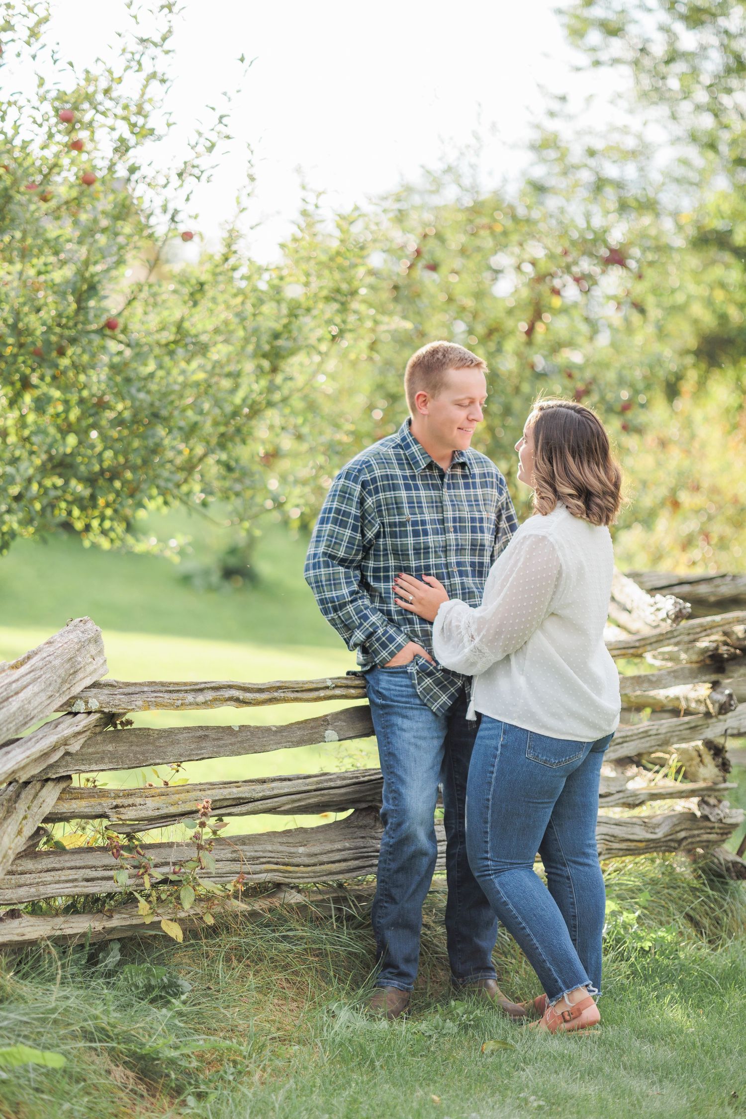 Couple embracing and smiling at each other beside a rustic wooden fence in apple orchard.