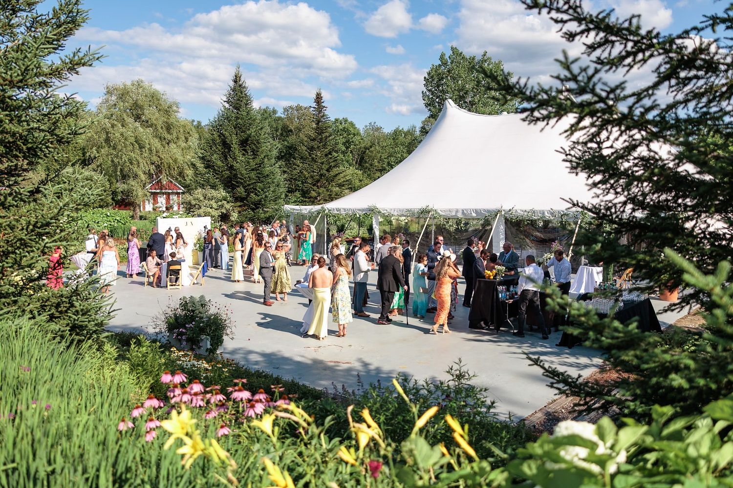 Wedding guests mingle under white tent at outdoor garden wedding reception on sunny summer day.