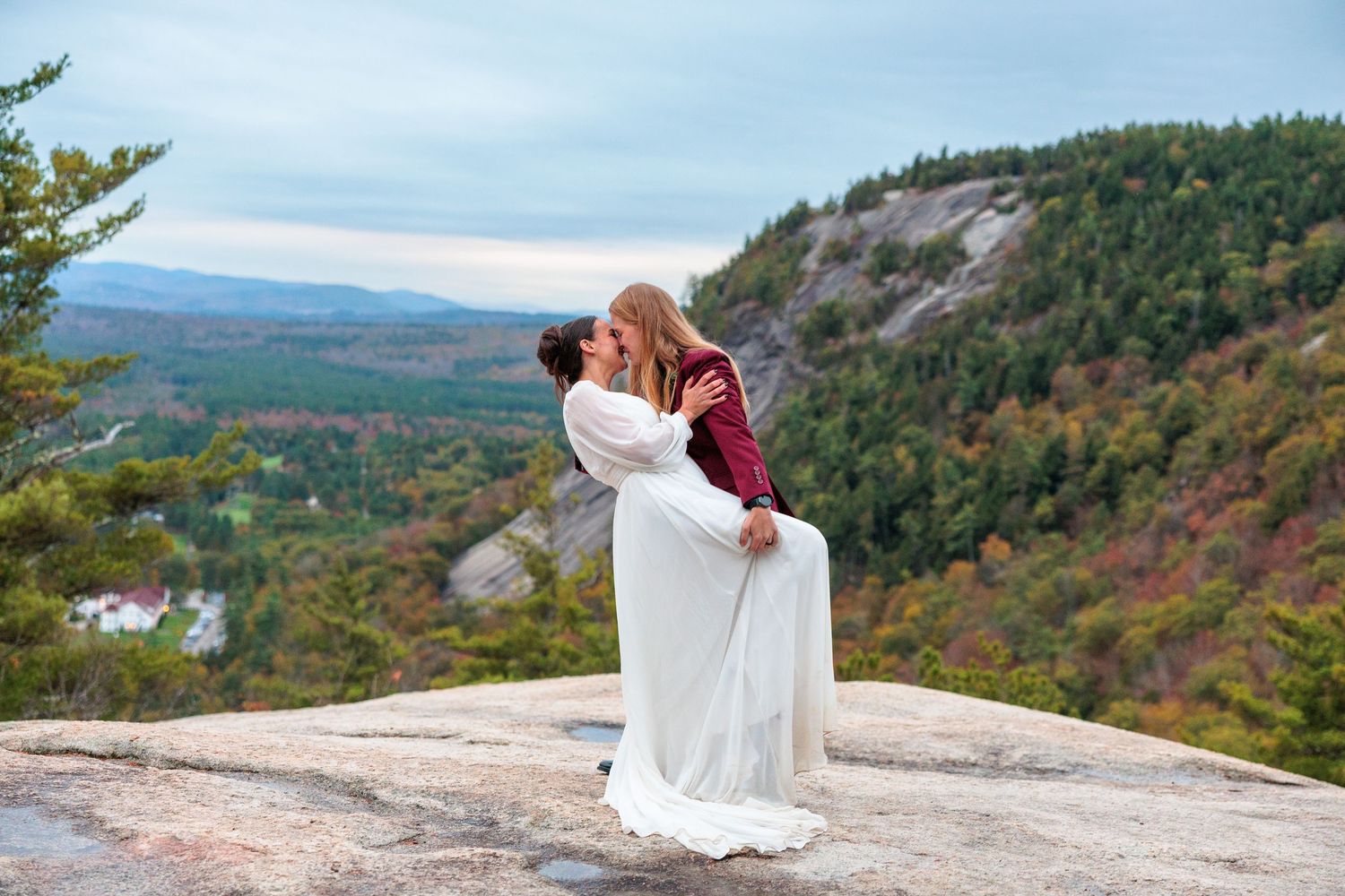 Couple sharing a romantic kiss on a mountain summit, bride in white gown lifted by partner, scenic valley backdrop.