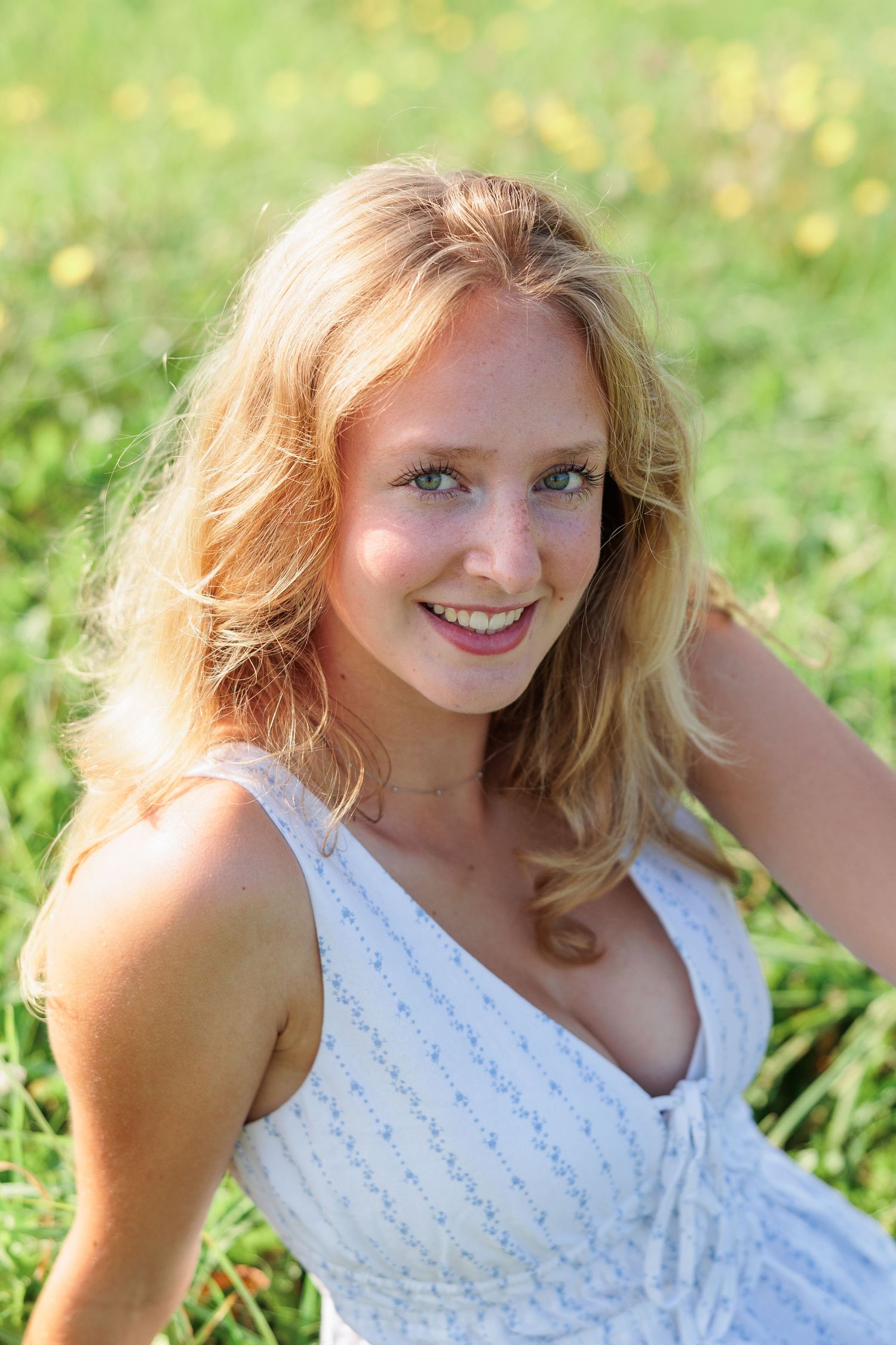 Smiling blonde woman with blue eyes sitting in green meadow wearing white sundress on sunny day.