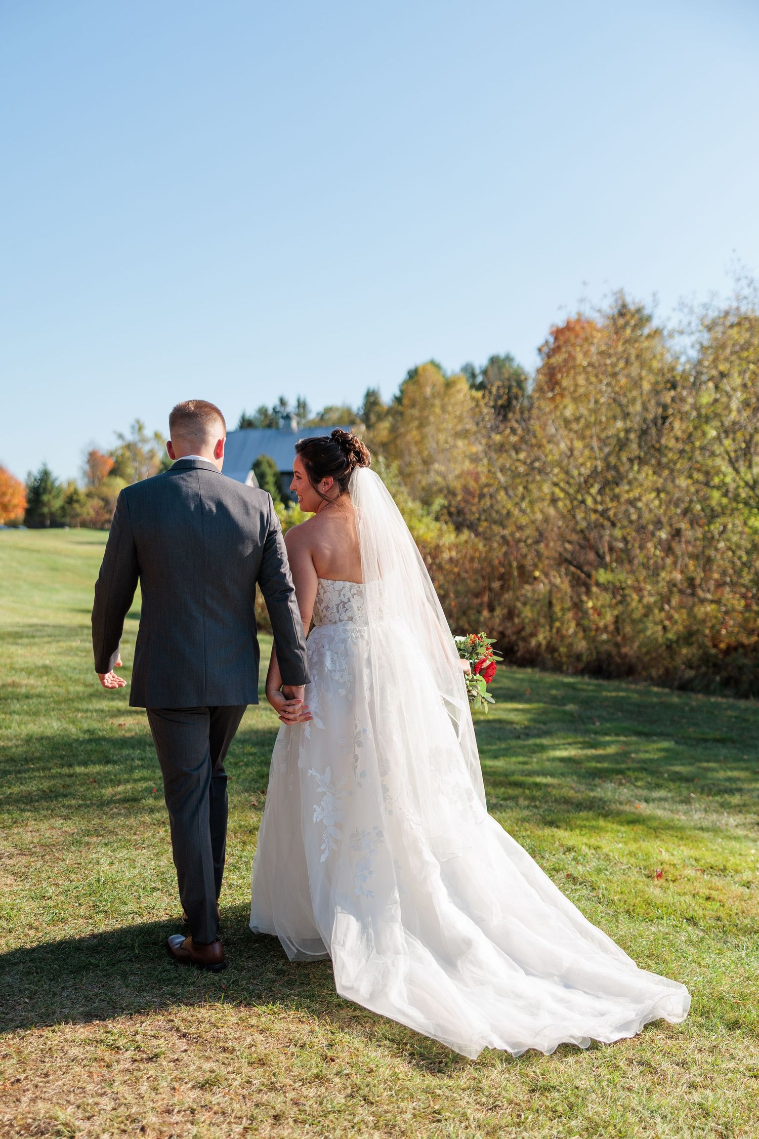 Bride and groom walking hand in hand across green lawn with colorful autumn foliage.