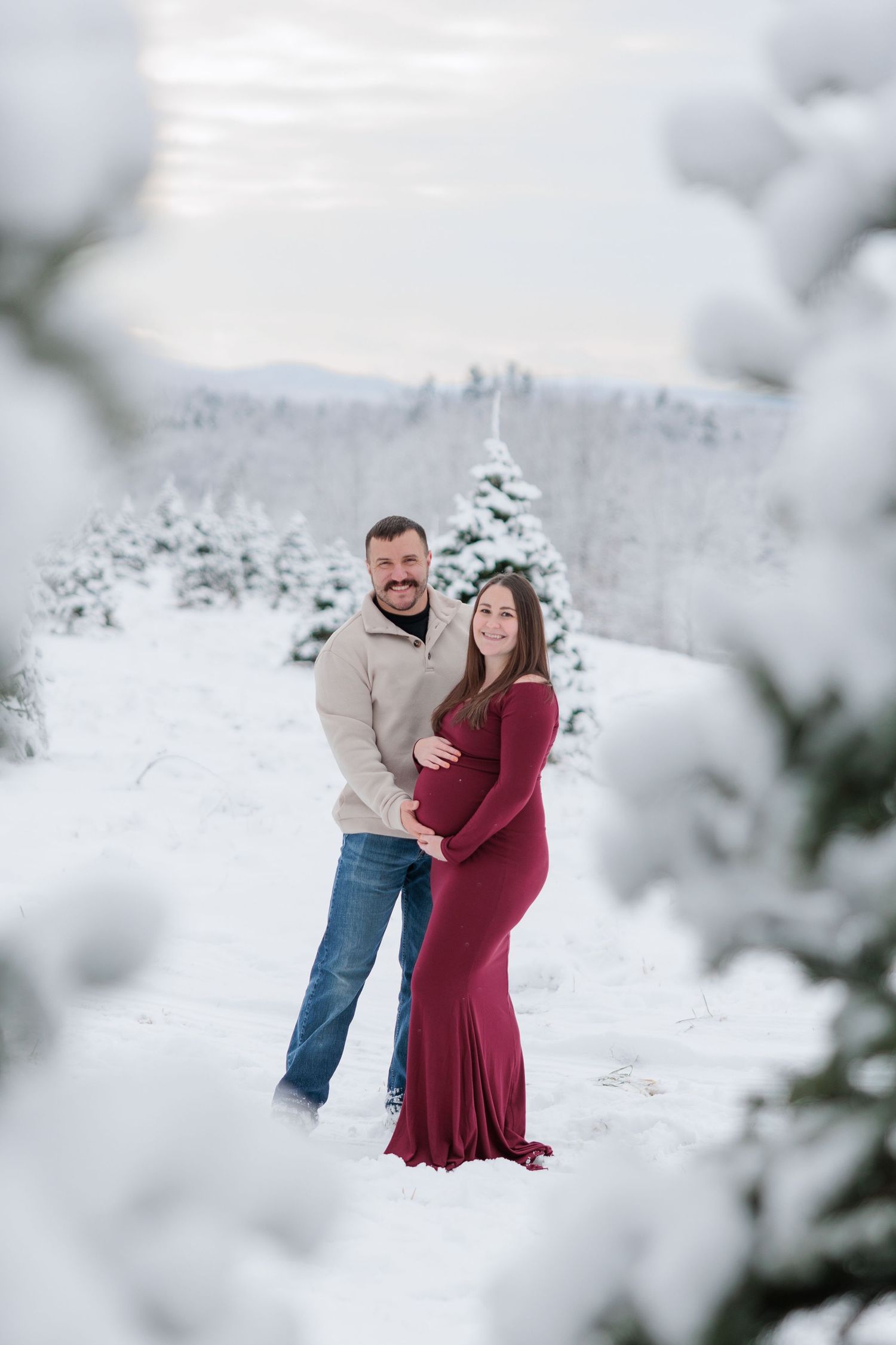 A couple poses together in a snowy winter landscape, the woman wearing a fitted burgundy maternity gown.