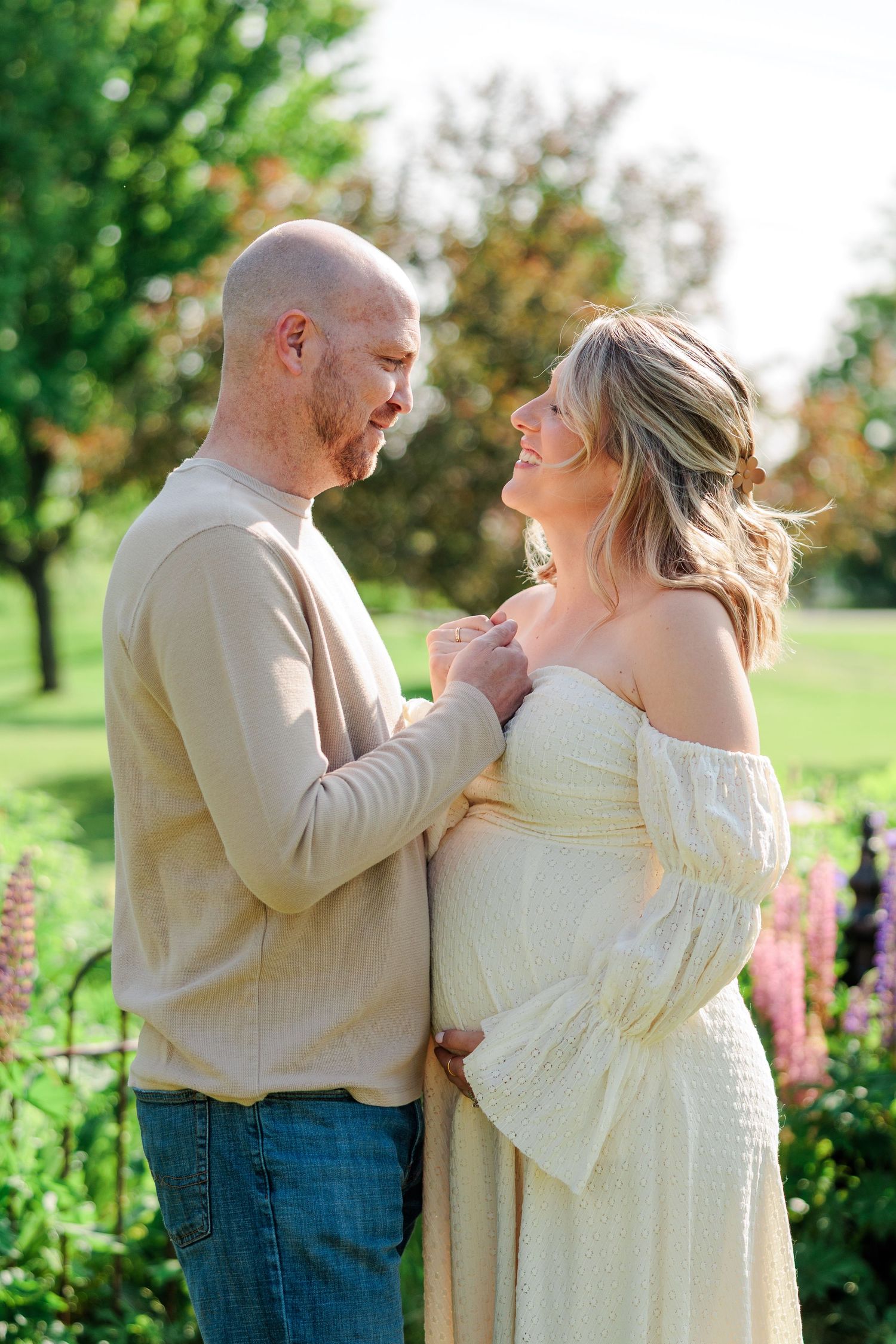 Bald man tenderly holding hands with smiling pregnant woman in white dress during outdoor maternity shoot.