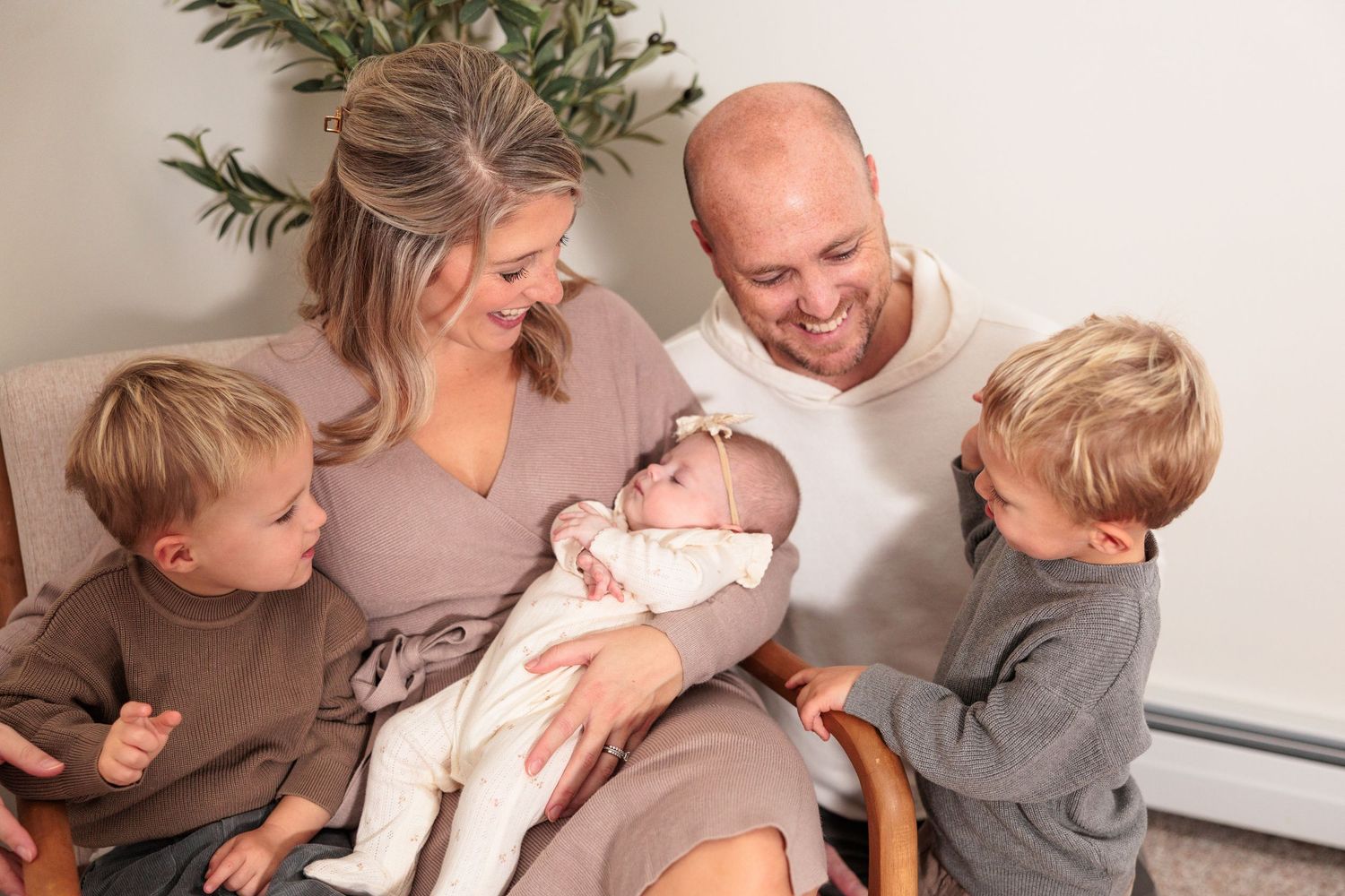 Happy family of four with parents, two young boys, and newborn baby girl gathered together smiling indoors.
