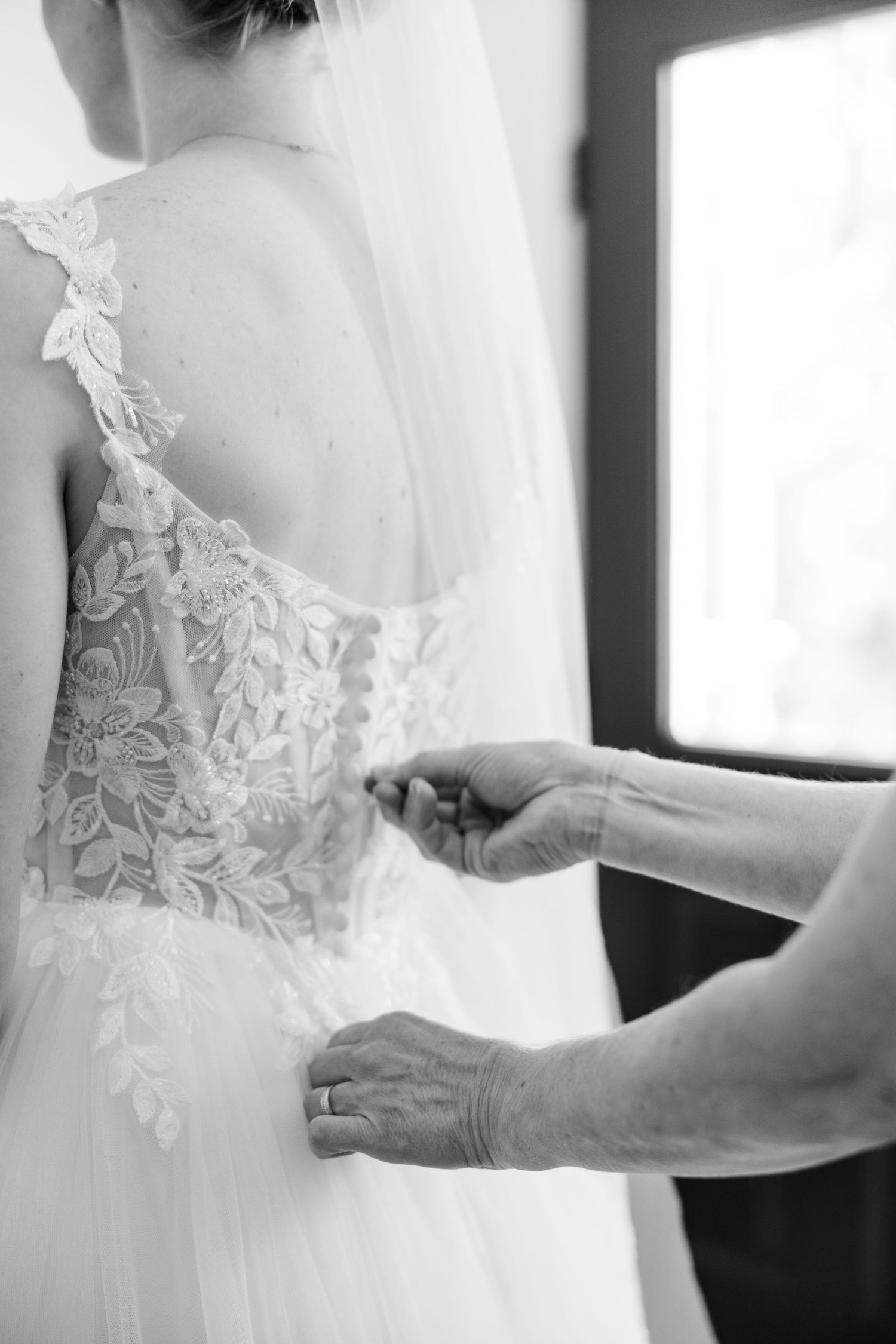 Black and white photo of hands fastening buttons on a bride's intricate lace wedding gown.