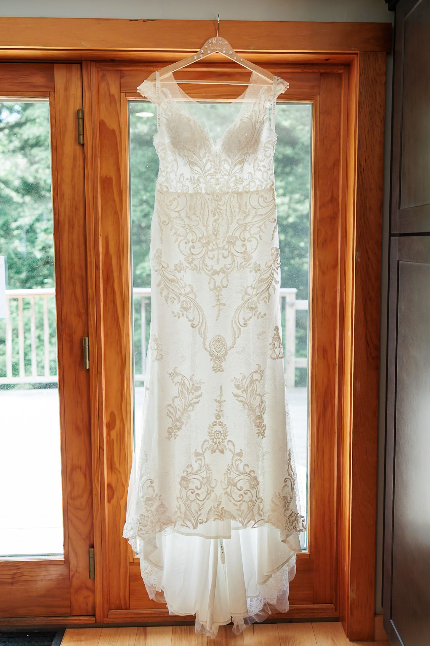 Elegant white lace wedding gown with gold embroidery hanging on wooden French doors before ceremony.