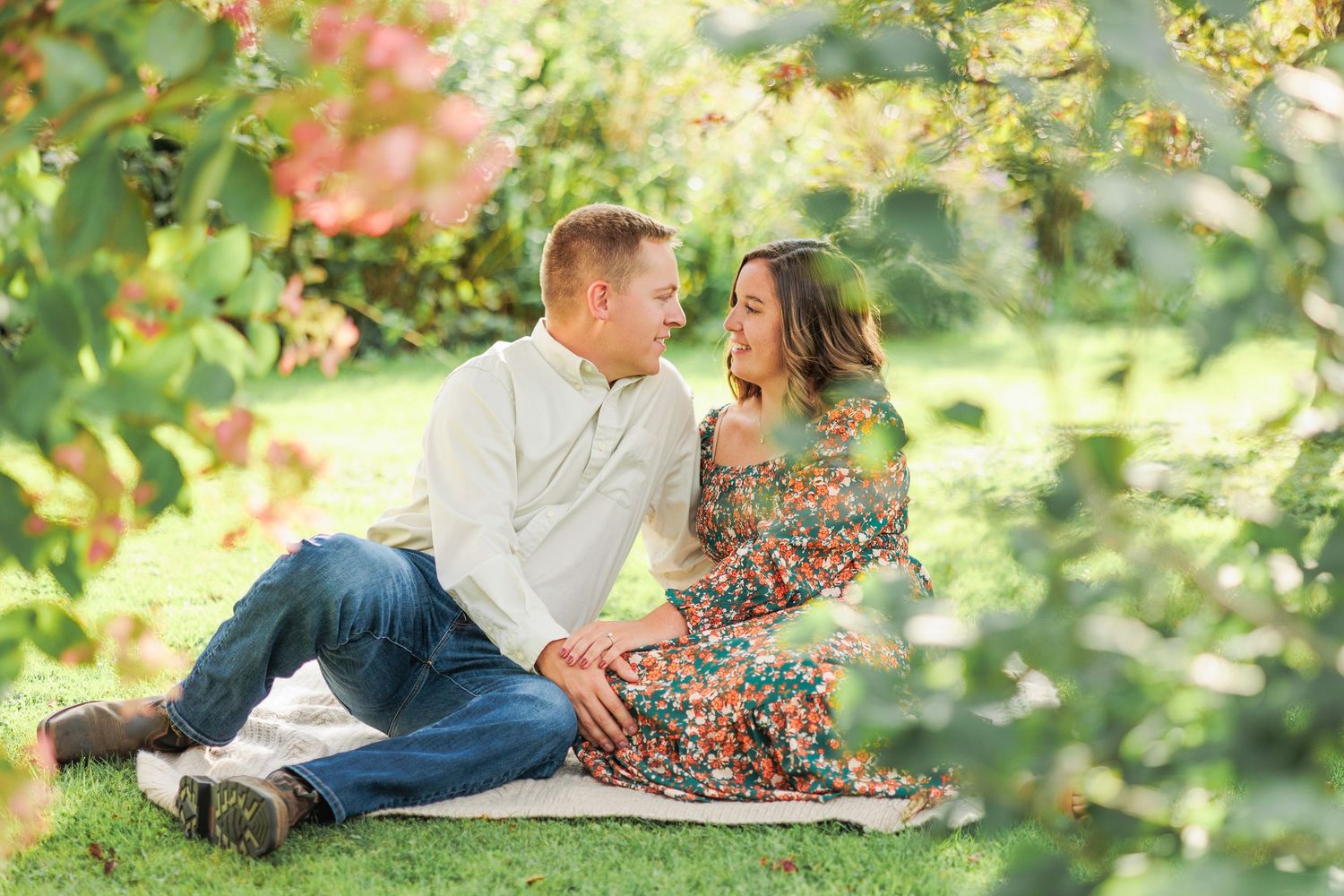Romantic couple gazing at each other while sitting on grass framed by lush green foliage.
