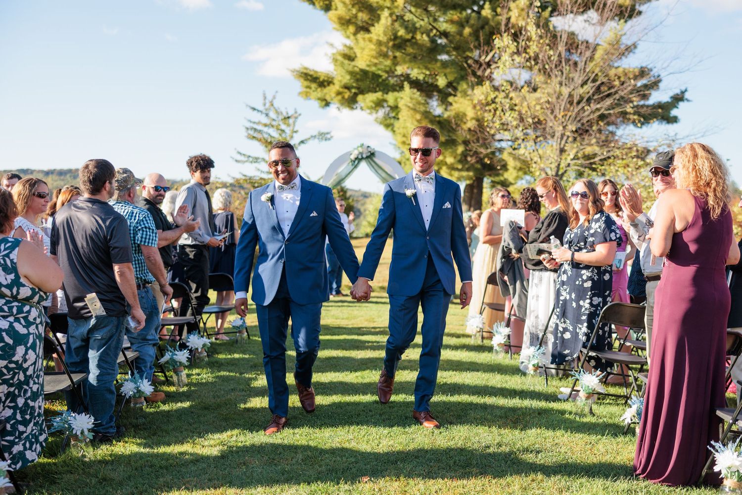 Wedding guests gather on lawn chairs during an outdoor ceremony with decorative white arch.