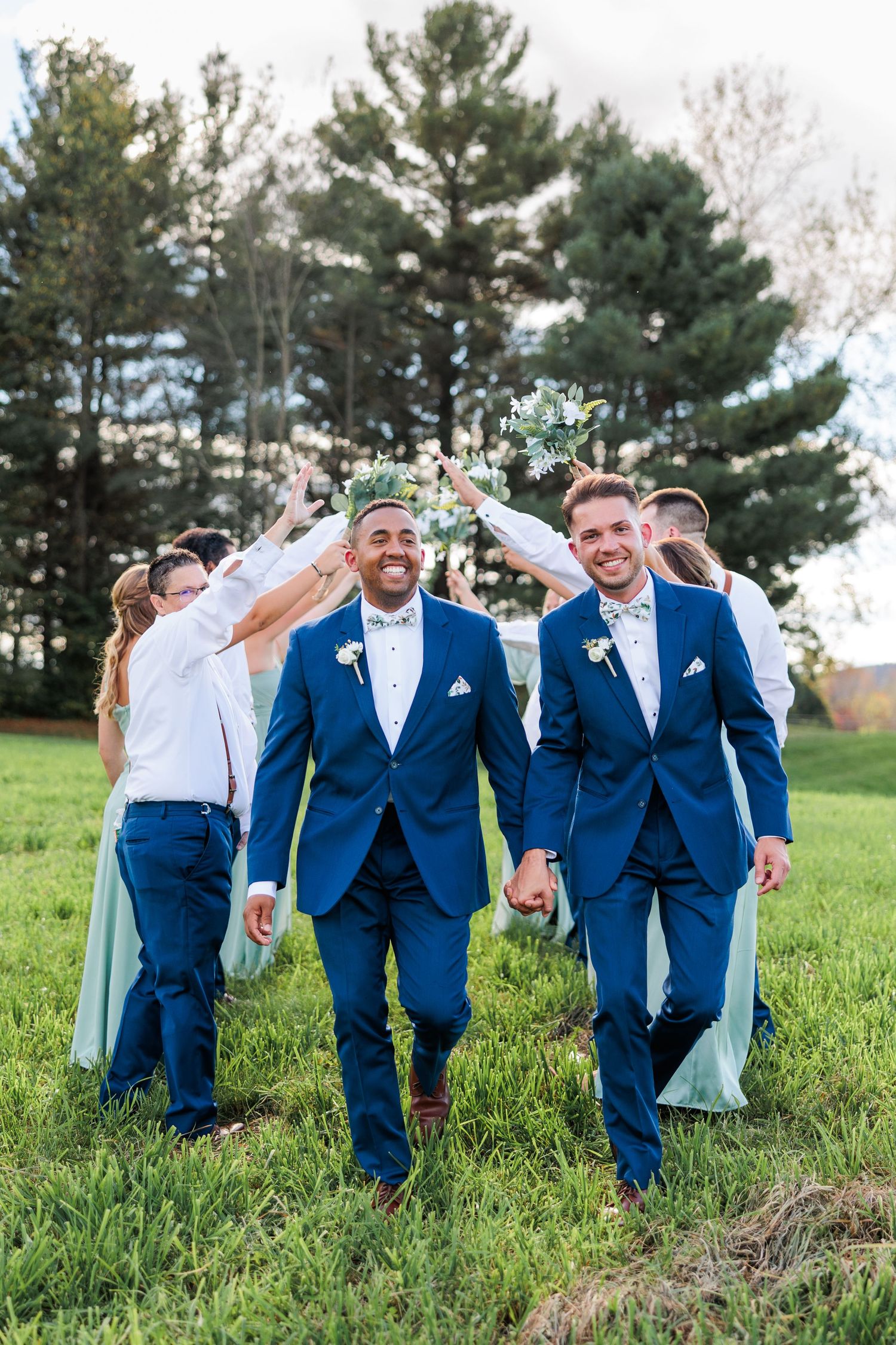 Wedding party members celebrate joyfully while walking across sunny grass field.