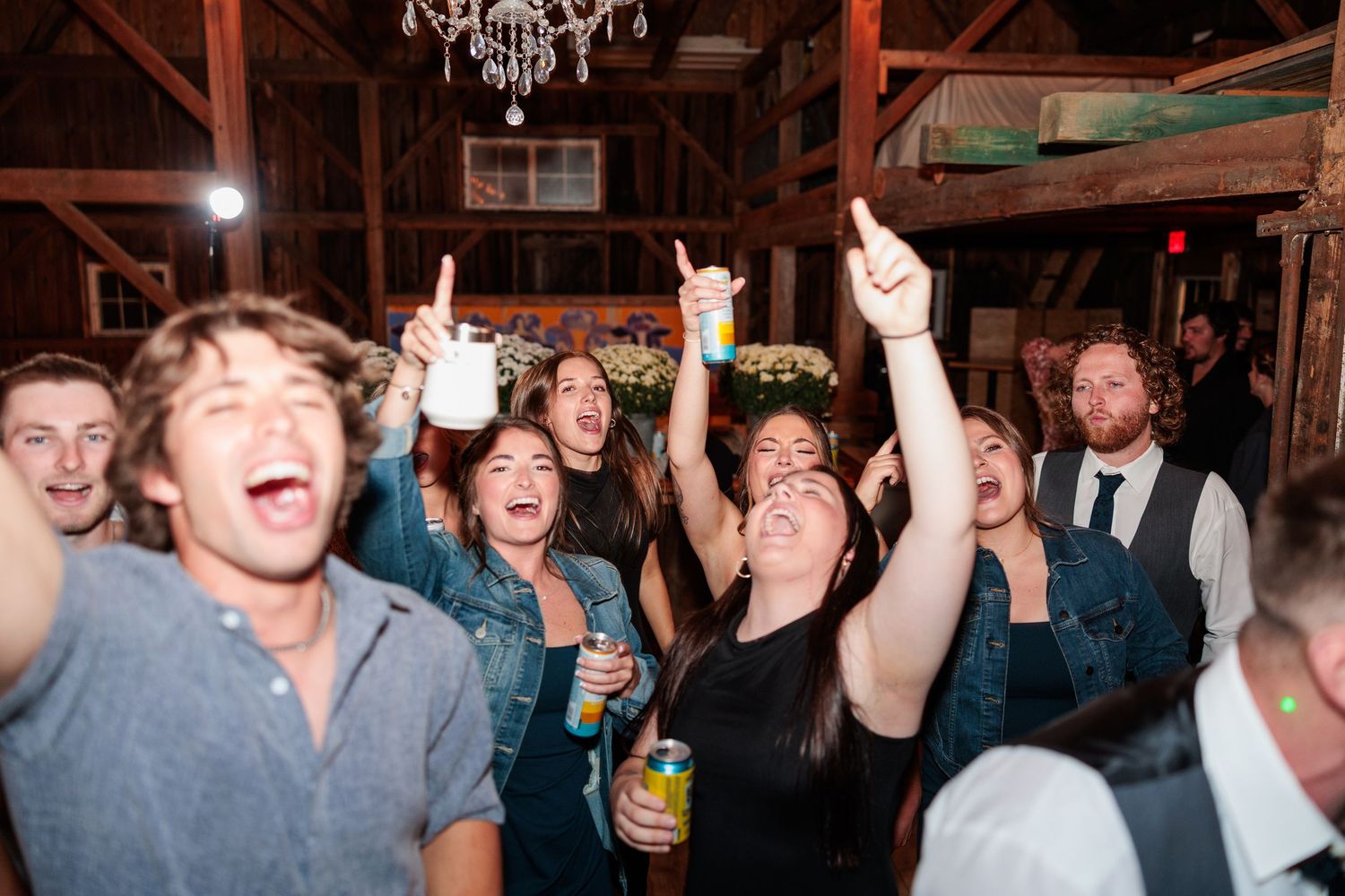 Wedding reception guests celebrate and dance inside a rustic barn venue with chandelier lighting.