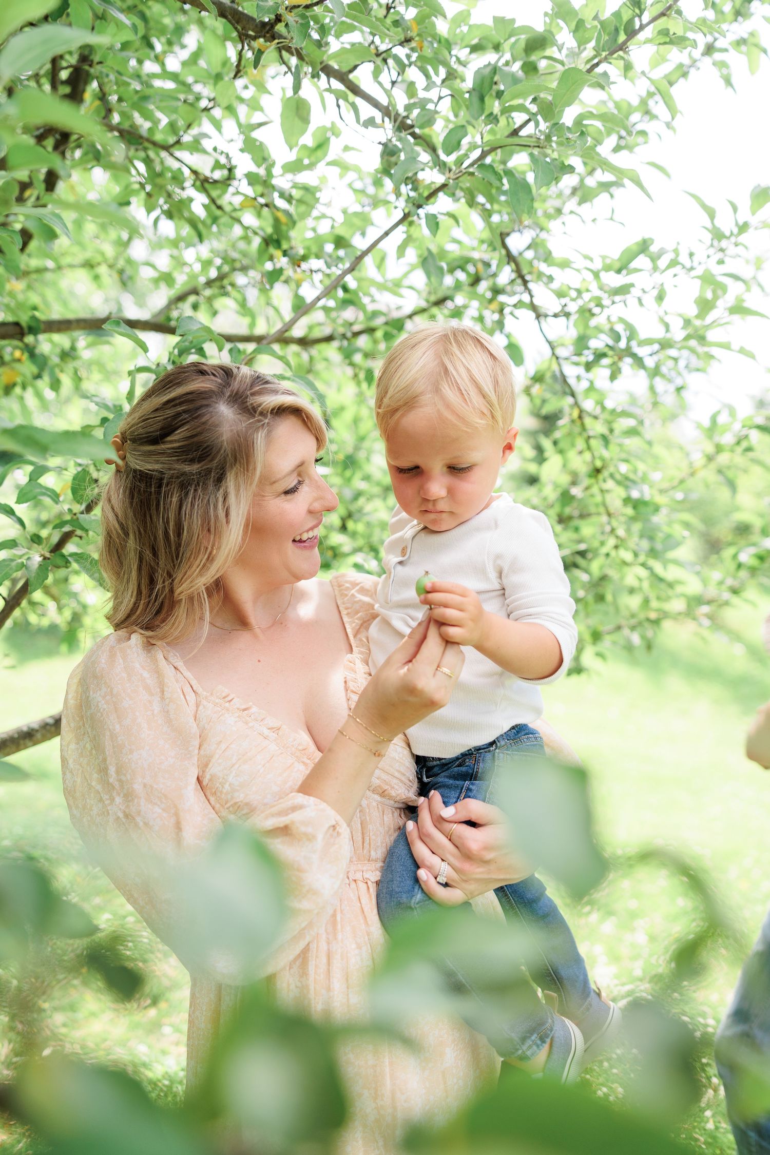 Mother holding toddler son under green apple tree branches in sunny garden, both examining small fruit.