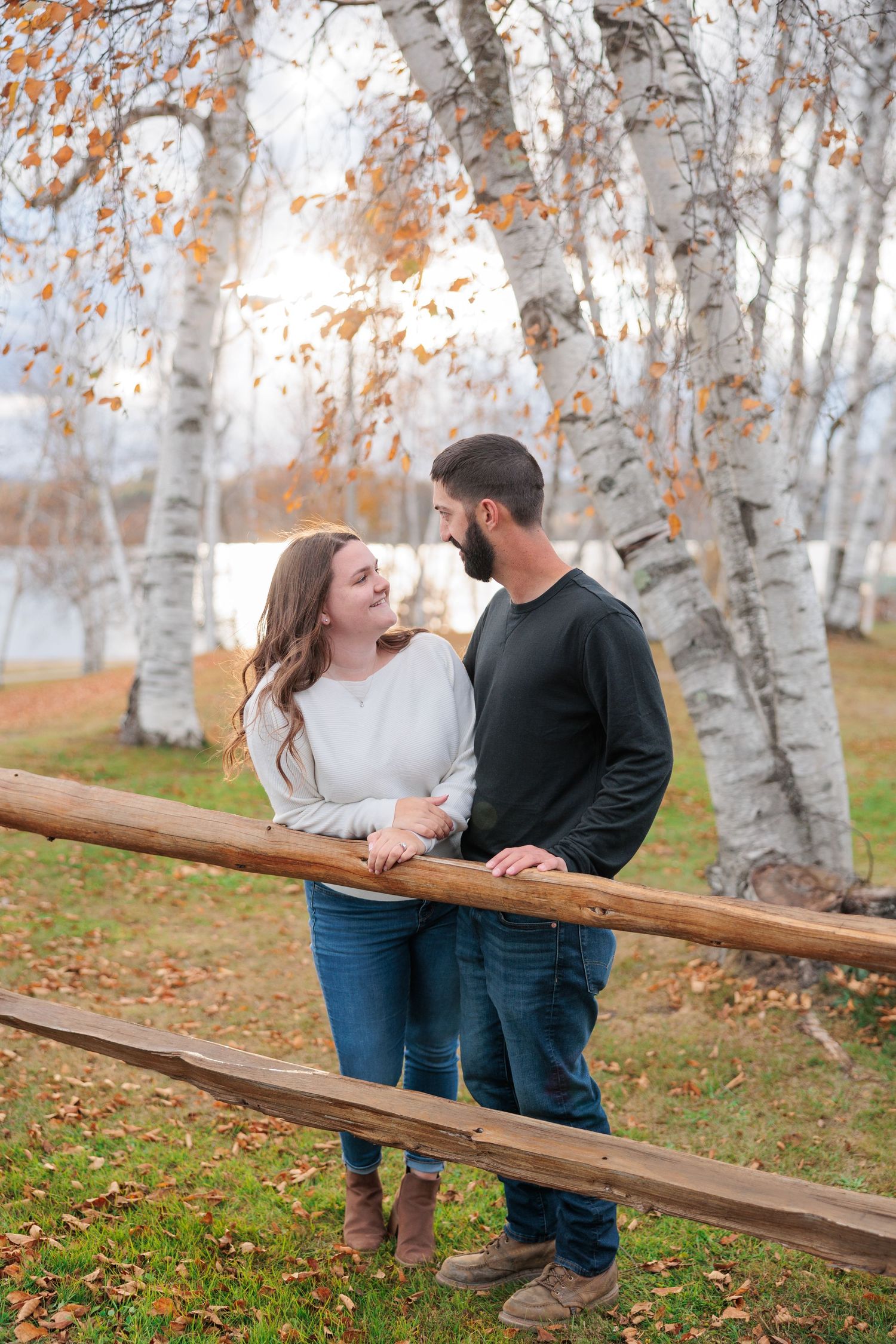 Couple smiling at each other while leaning on a rustic wooden fence surrounded by white birch trees in fall.