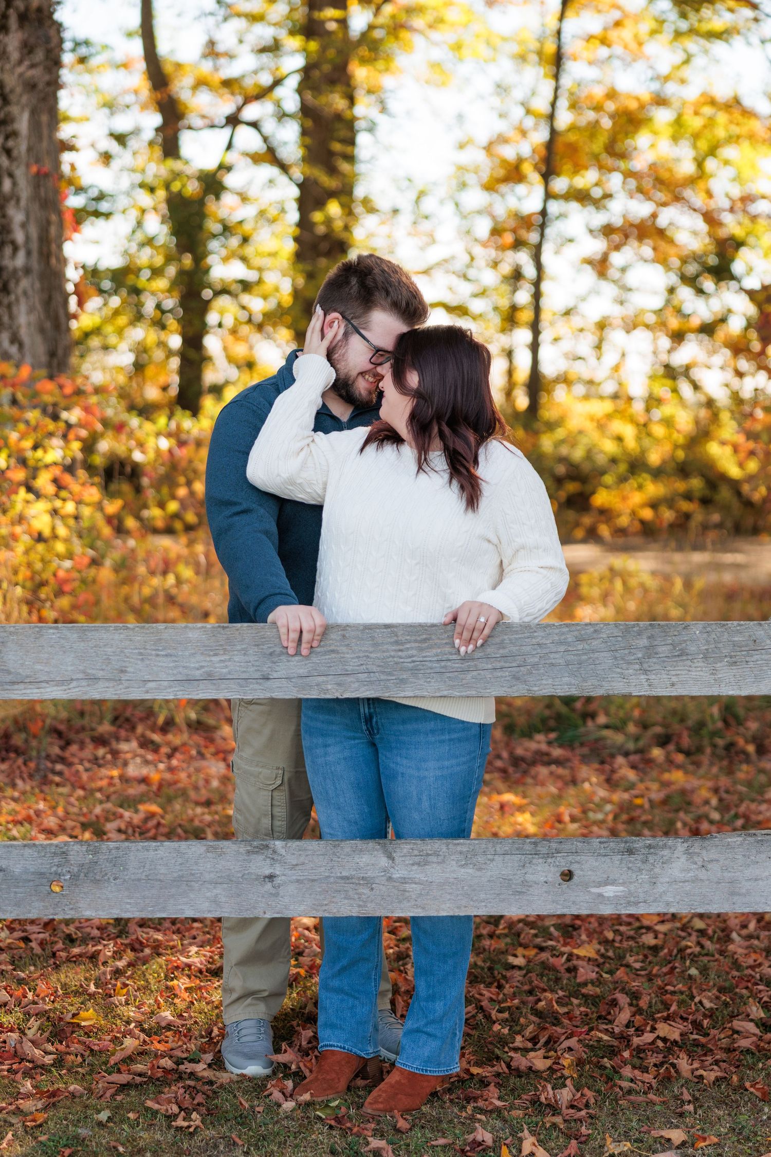 Couple sharing a tender kiss leaning on a rustic wooden fence surrounded by colorful autumn foliage.