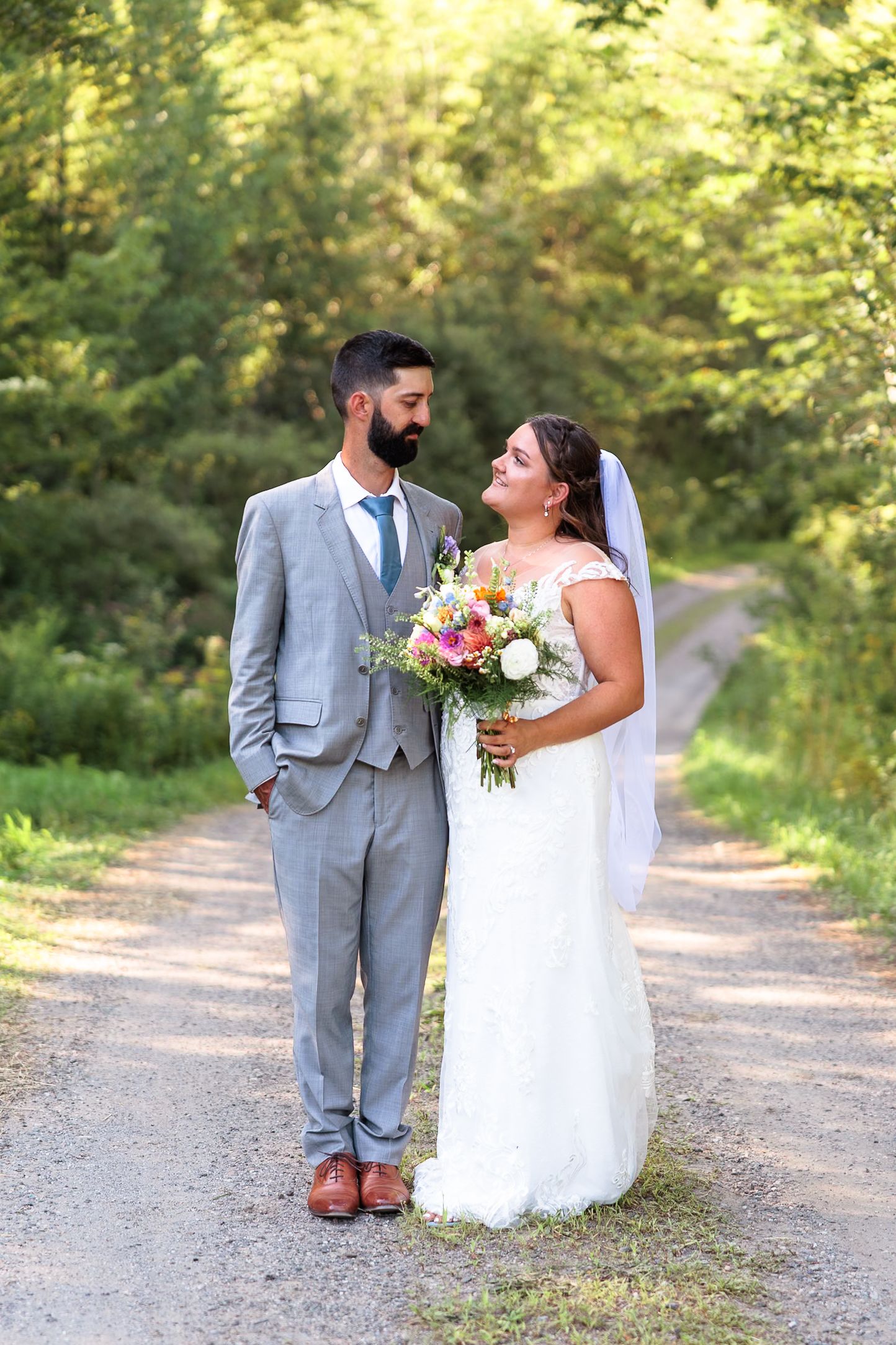 Bride and groom share a tender moment on a sunlit forest path, bride holding a colorful bouquet.