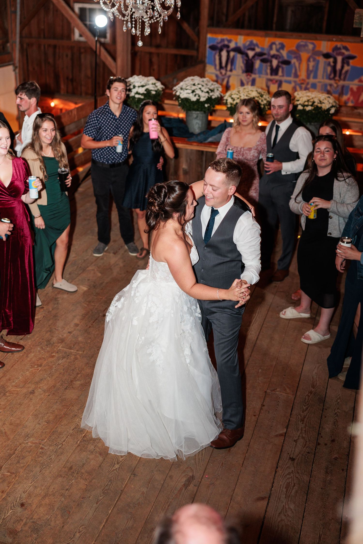 Bride and groom dance together in a warmly lit barn as wedding guests watch and celebrate.