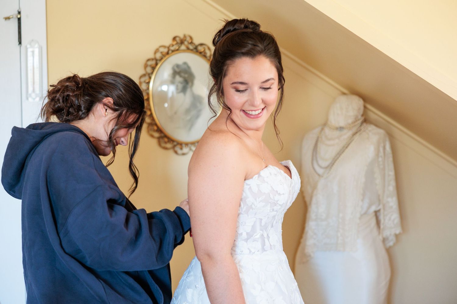 Bridesmaid helping bride zip up her lace wedding dress in a bright dressing room.
