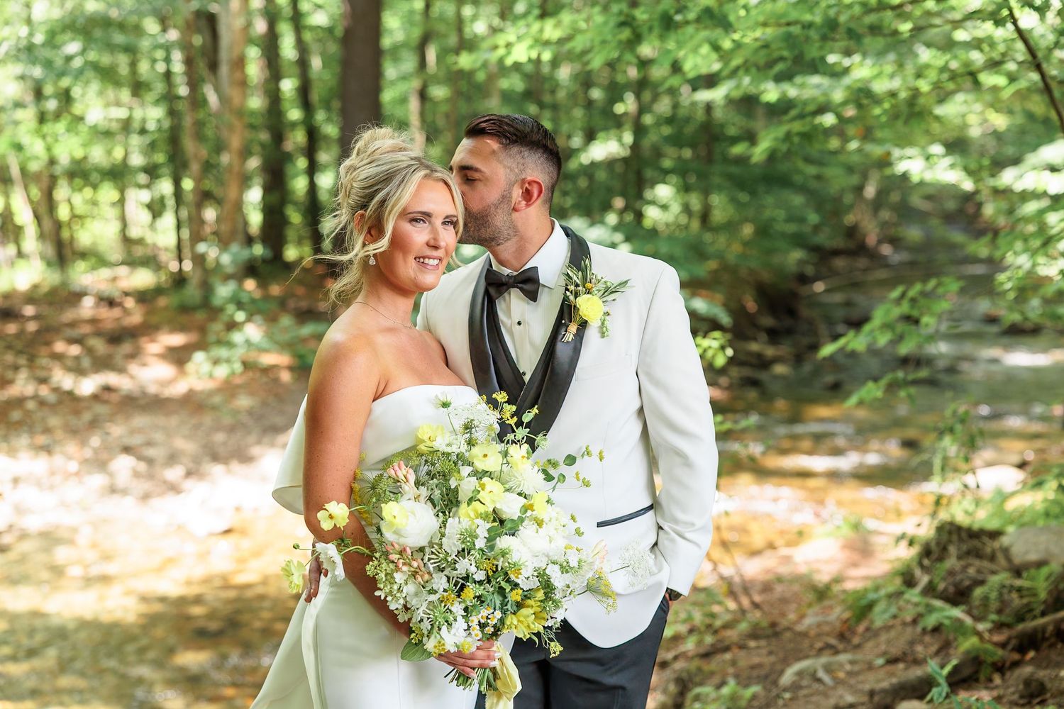 Happy bride and groom posing together in a sunlit forest, bride holding a white and yellow bouquet.