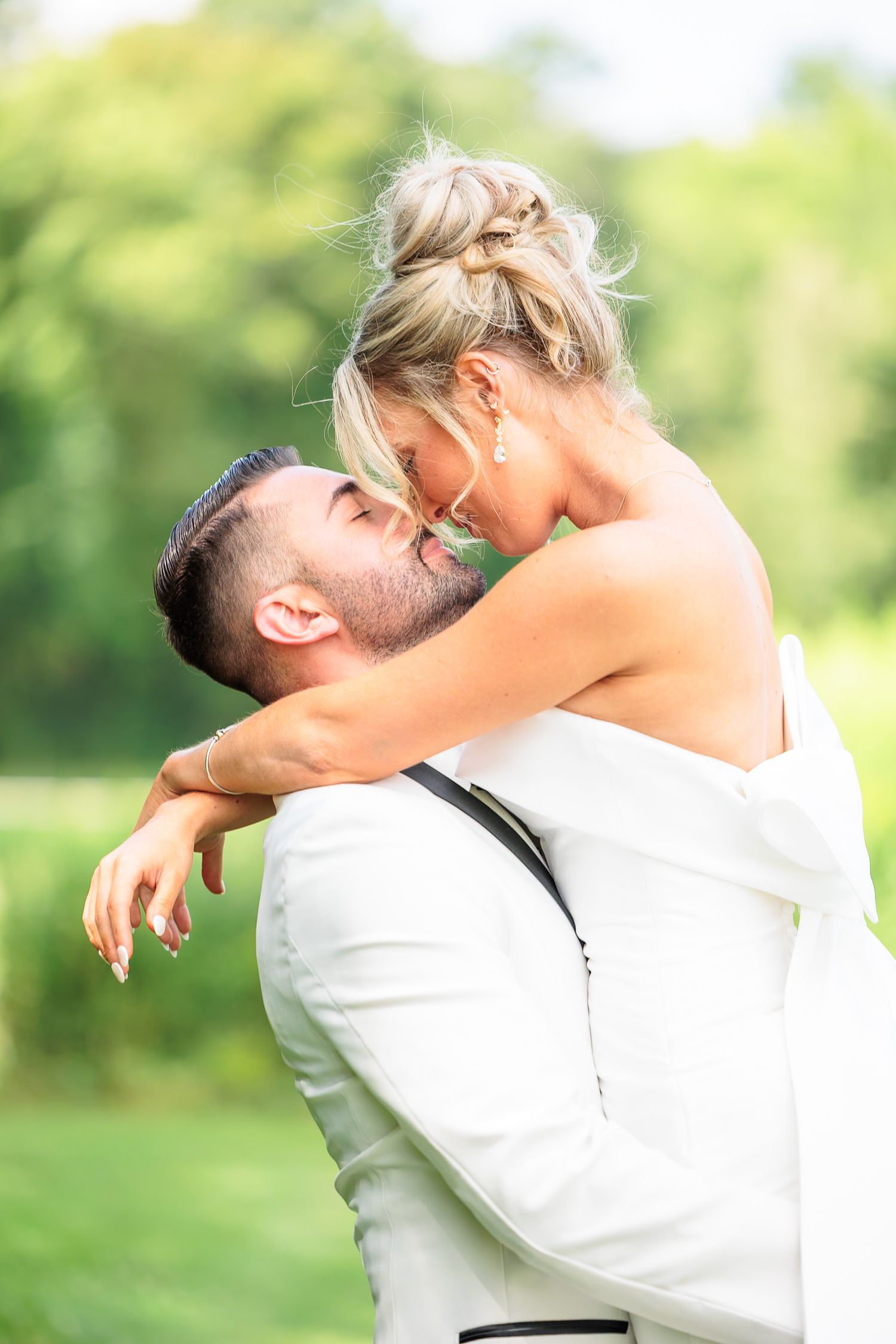 Groom lifts bride outdoors in lush green setting, sharing a romantic moment in matching white wedding attire.