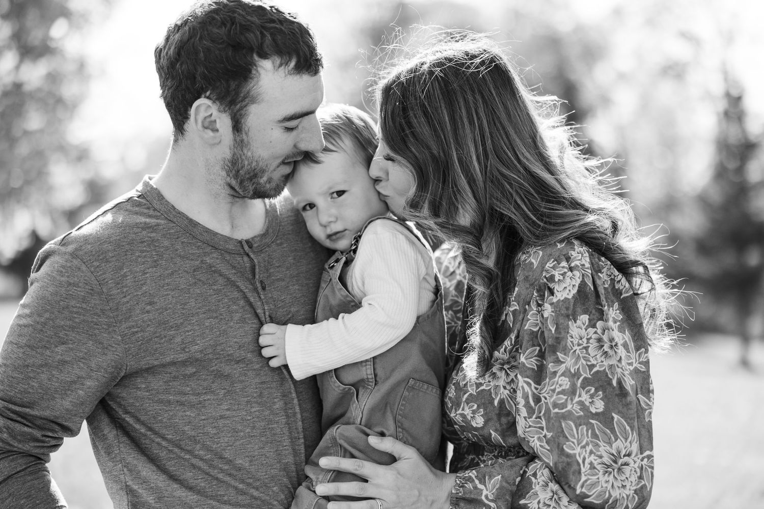 Black and white photo of parents lovingly kissing their baby on cheeks outdoors in nature.