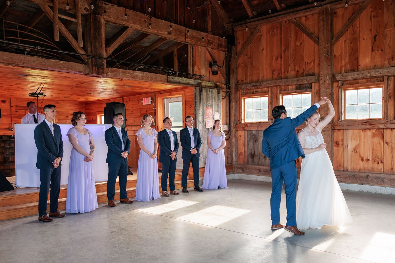 Wedding party watches as bride and groom share their first dance in a sunlit rustic barn.