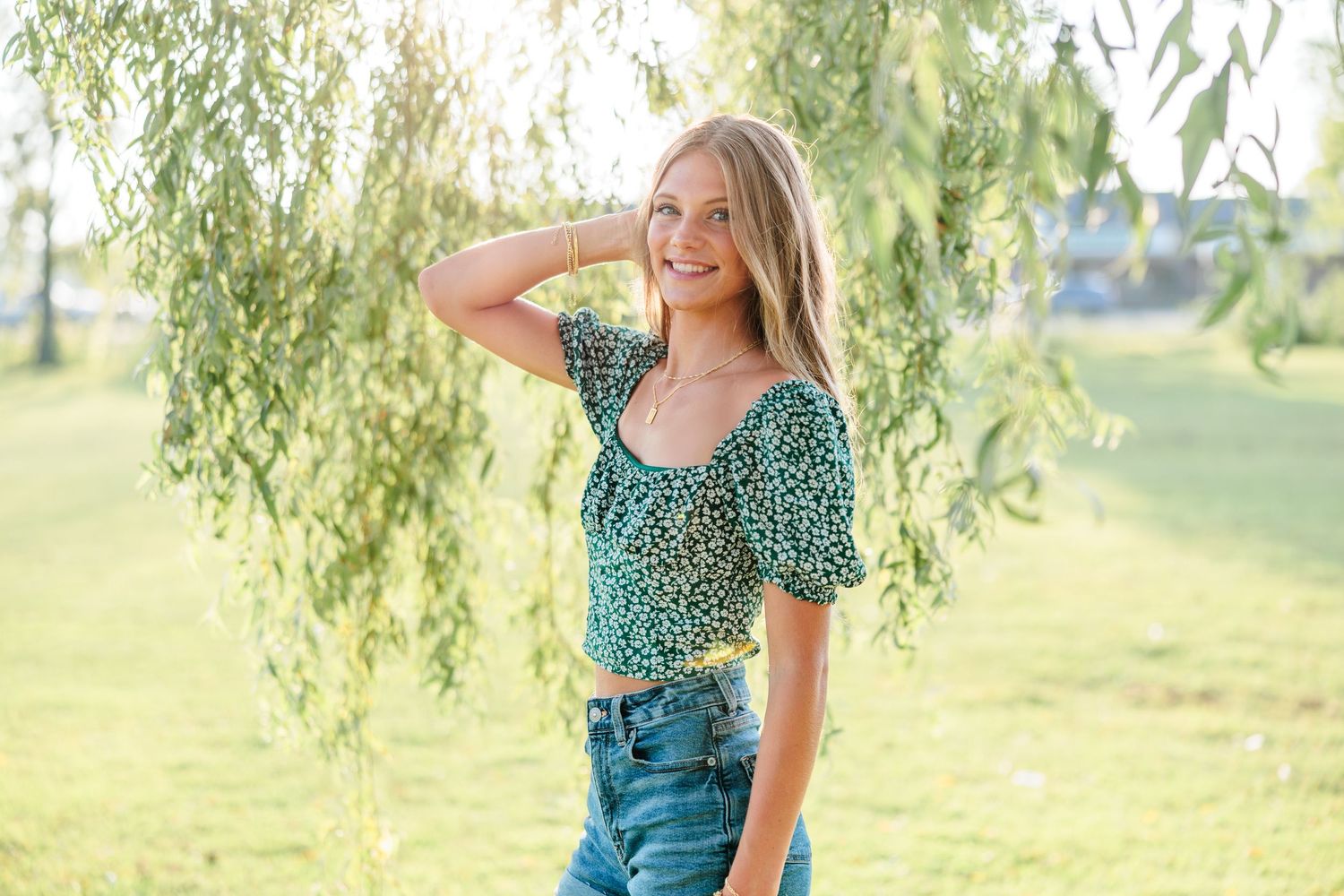 Smiling young woman in green floral crop top and jeans posing outdoors near willow tree on sunny day.