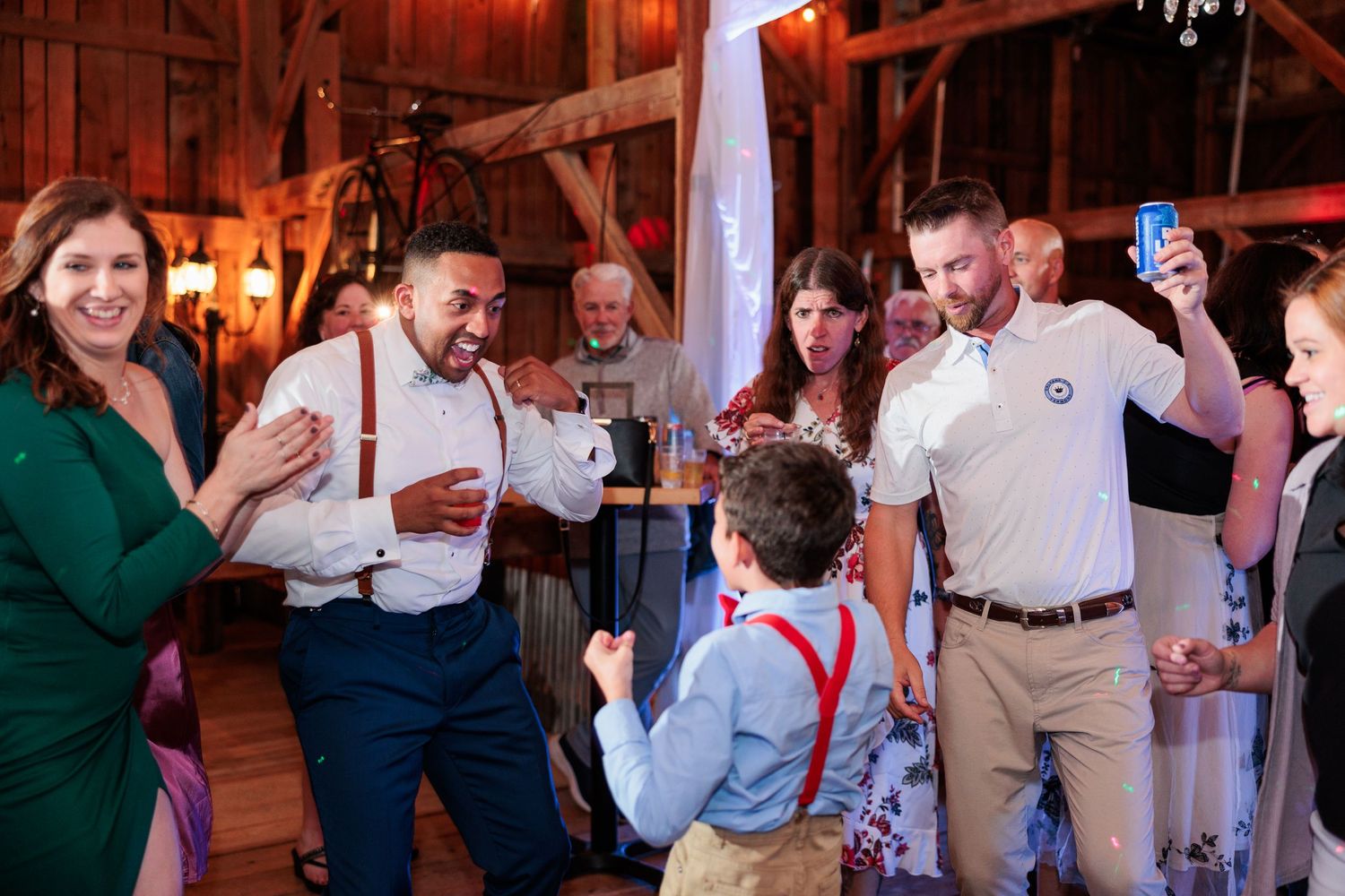 Wedding reception guests dancing and celebrating inside a rustic wooden barn with chandeliers.