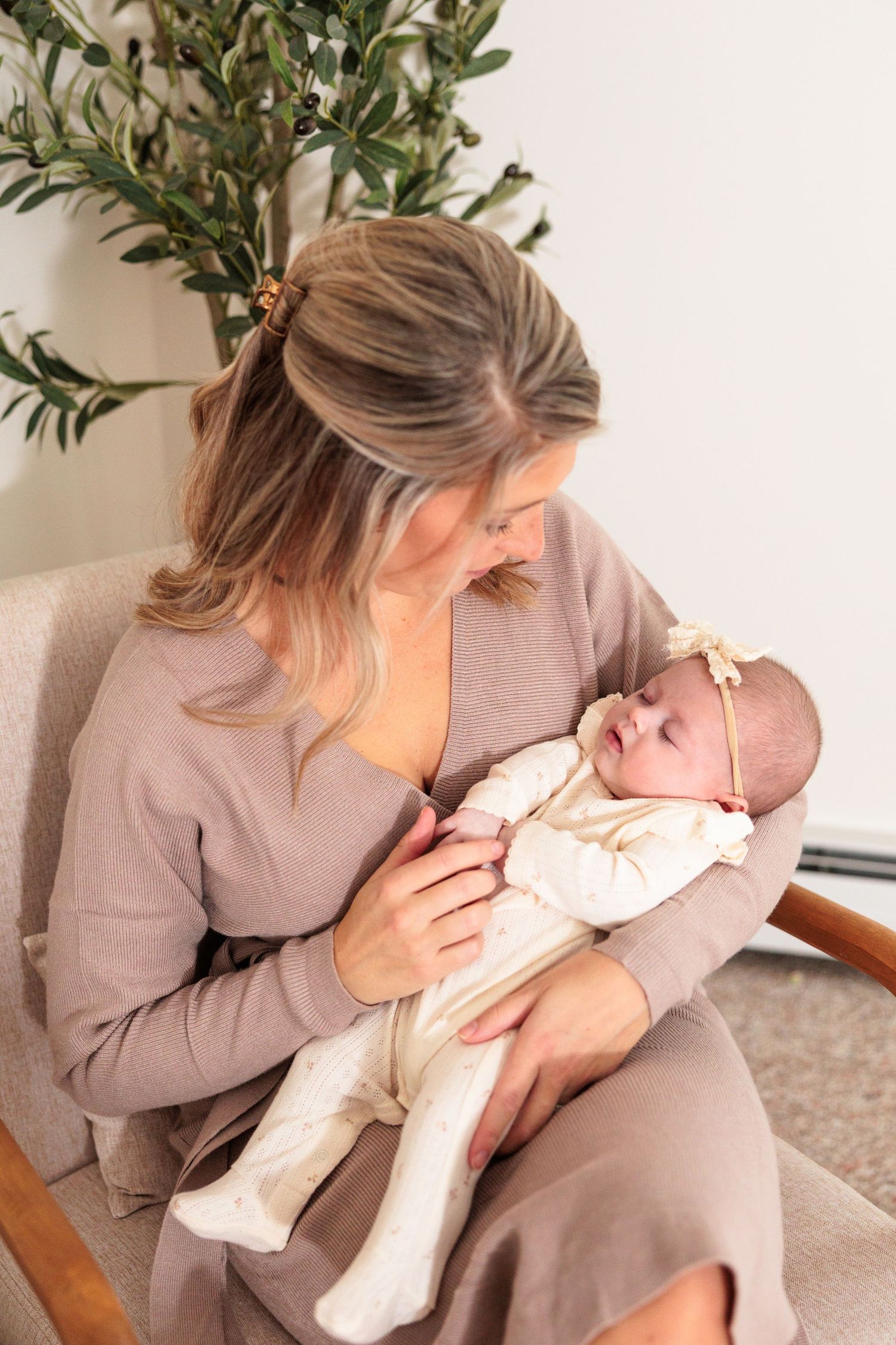 Mother in beige sweater holding sleeping infant wearing white outfit and bow in nursery setting.