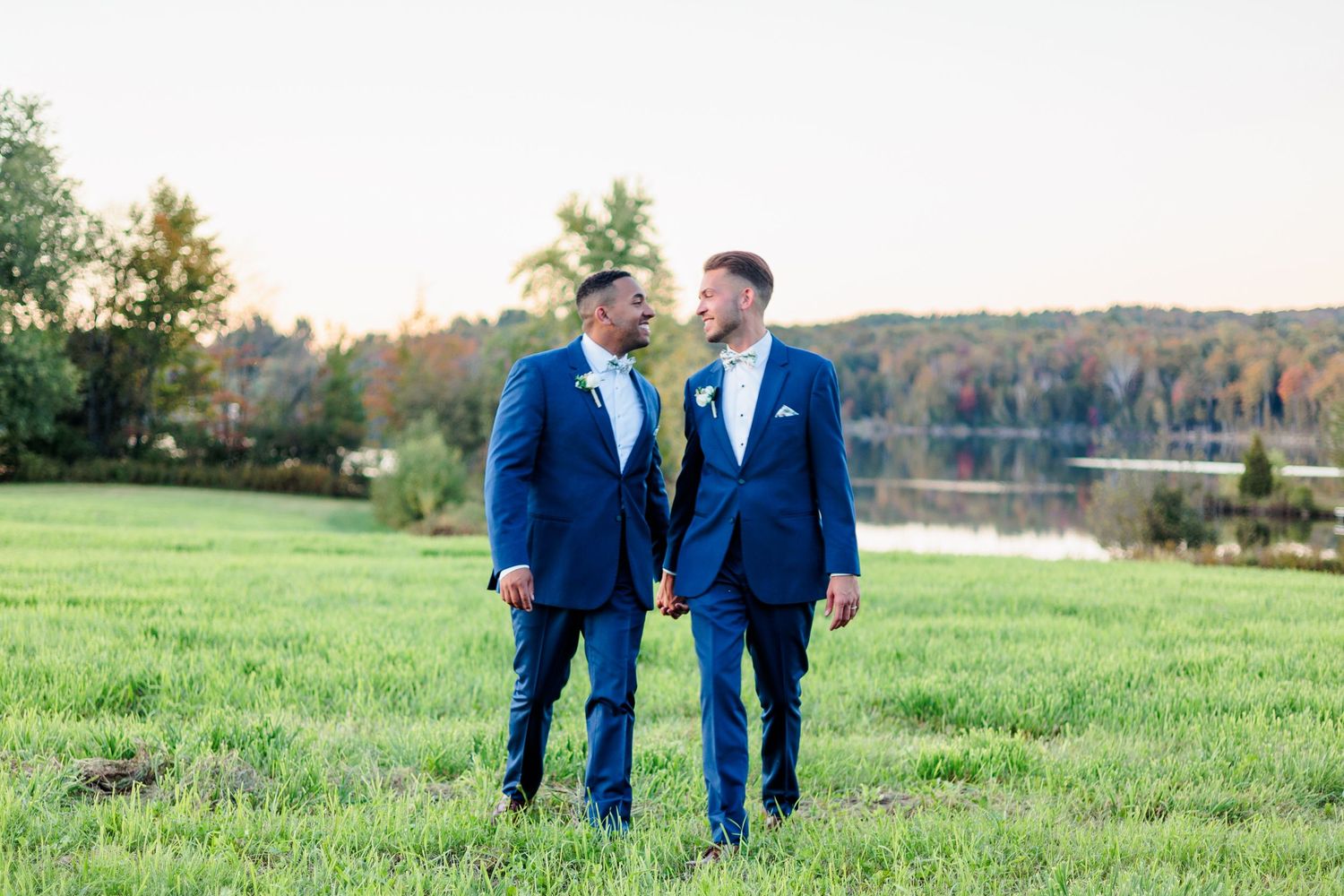 Wedding couple walking through green field with autumn trees in background.