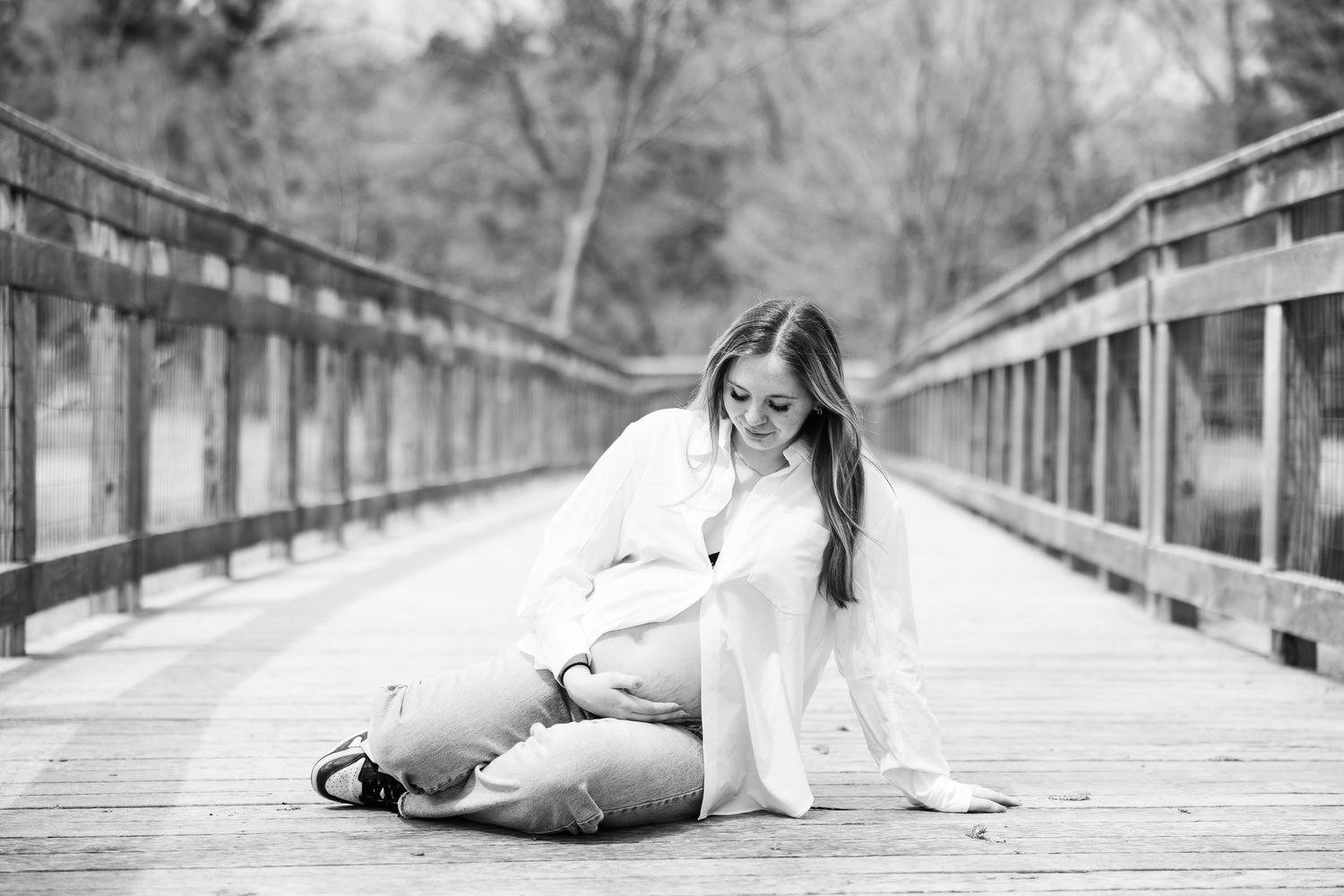 Black and white maternity photo of pregnant woman sitting on a wooden bridge deck outdoors.