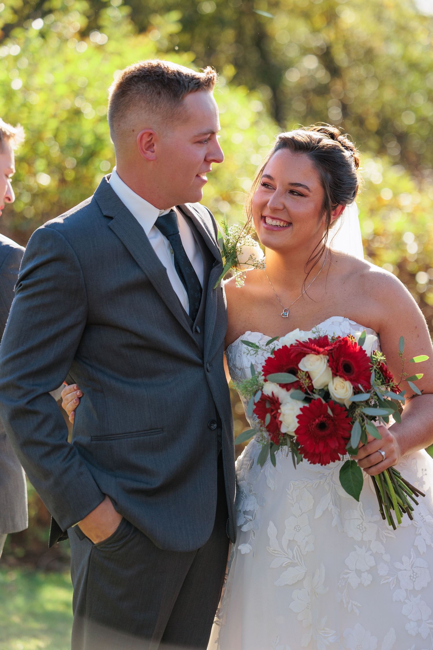 Smiling bride in strapless lace gown holding red bouquet stands with groom in gray suit at outdoor wedding.