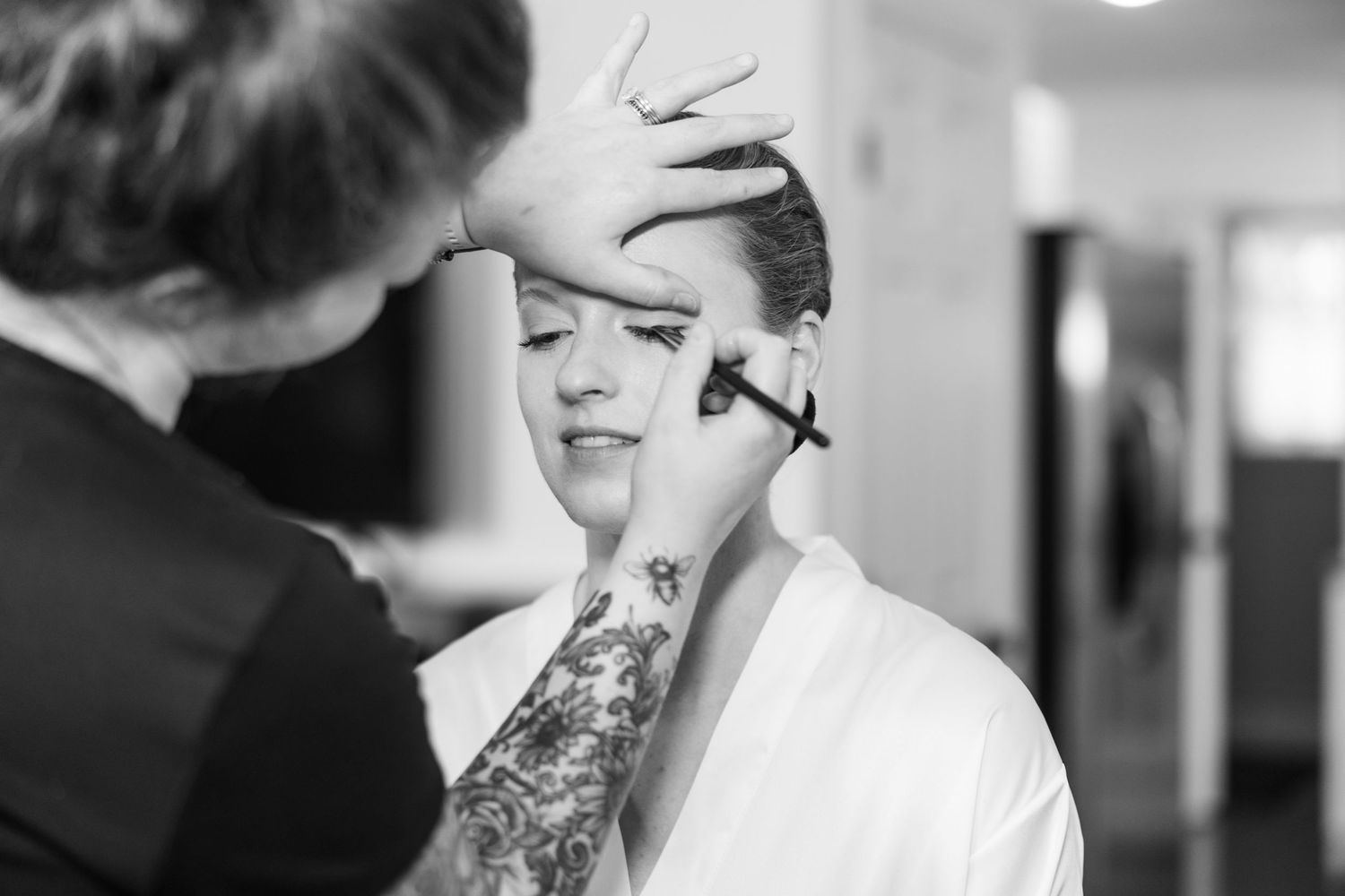 Black and white photo of a makeup artist applying foundation to a bride in a white robe.