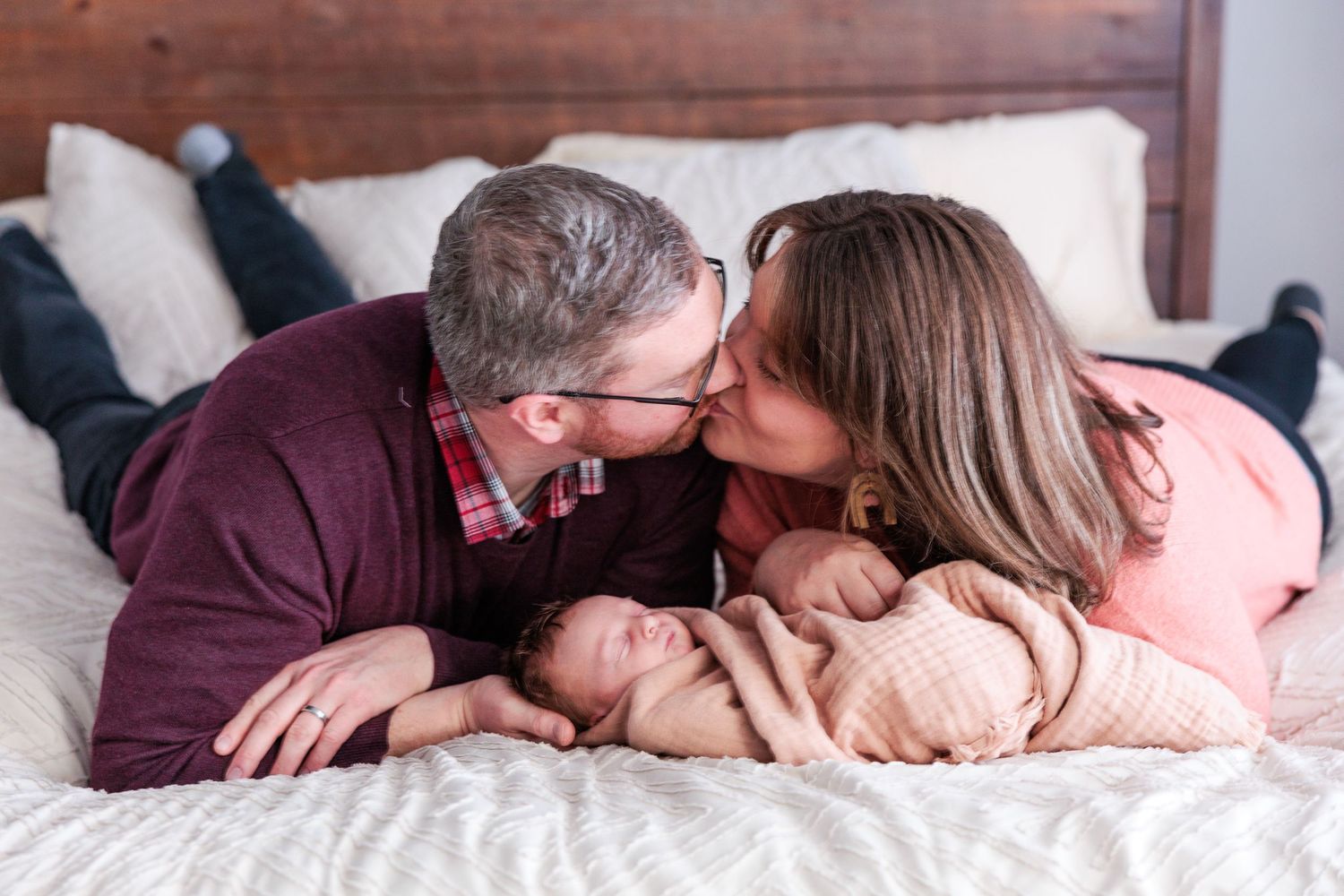 Parents kissing on bed while newborn baby rests between them on white bedding.