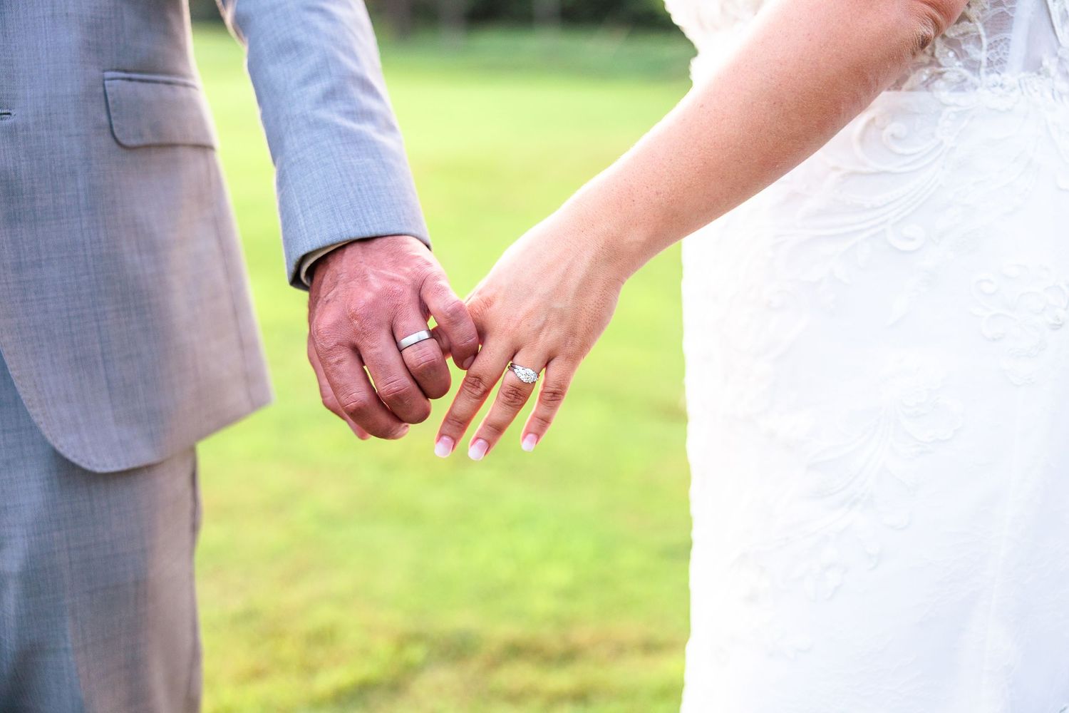 Close-up of bride and groom holding hands showing wedding rings in outdoor ceremony.
