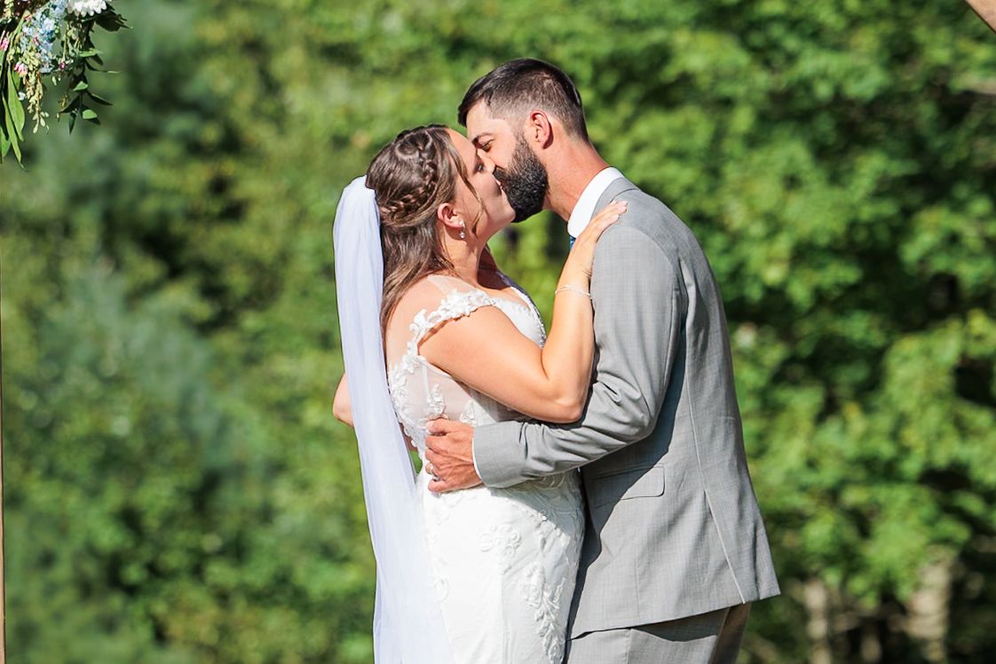Bride and groom share a romantic first kiss surrounded by lush green trees at outdoor wedding.