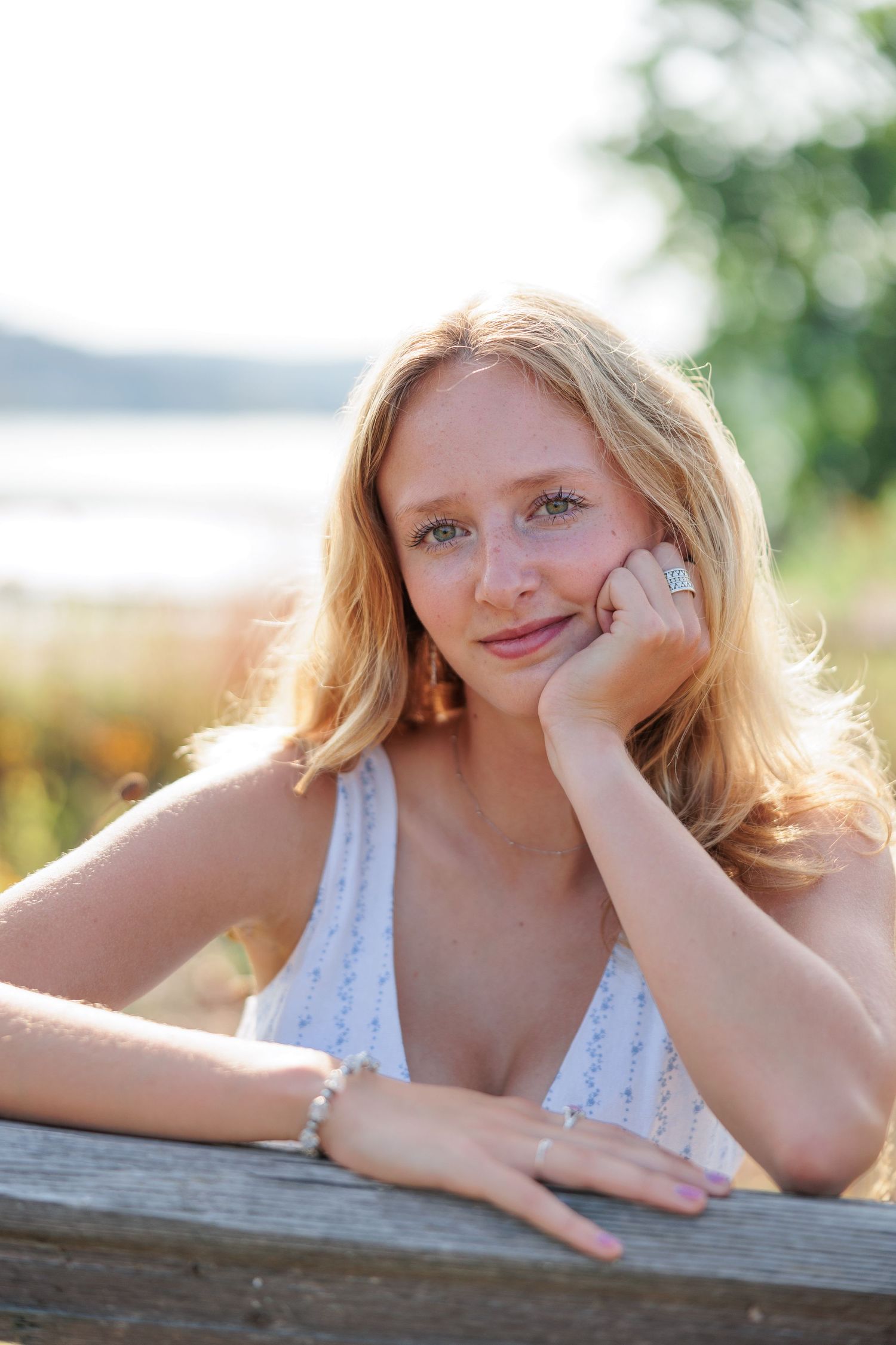 Blonde woman resting chin on hand at wooden fence outdoors near lake in golden sunlight.