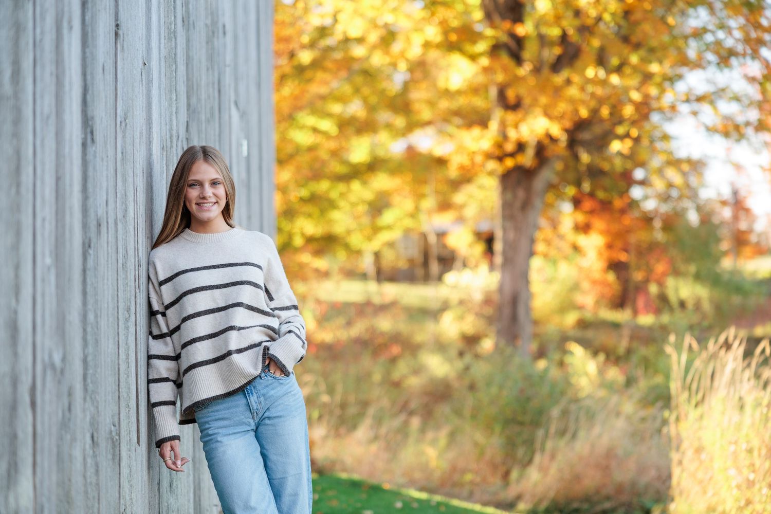Teen girl in striped sweater and jeans leaning against barn wall with colorful autumn foliage in background.
