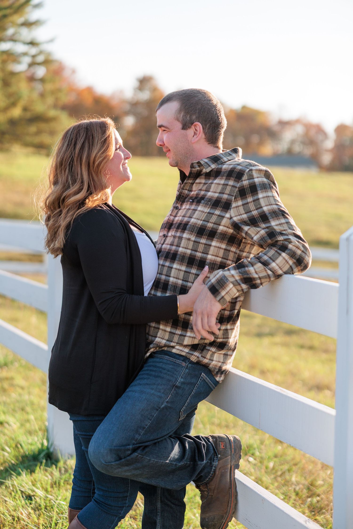 Engaged couple smiling at each other while leaning against a white fence in a sunny field.