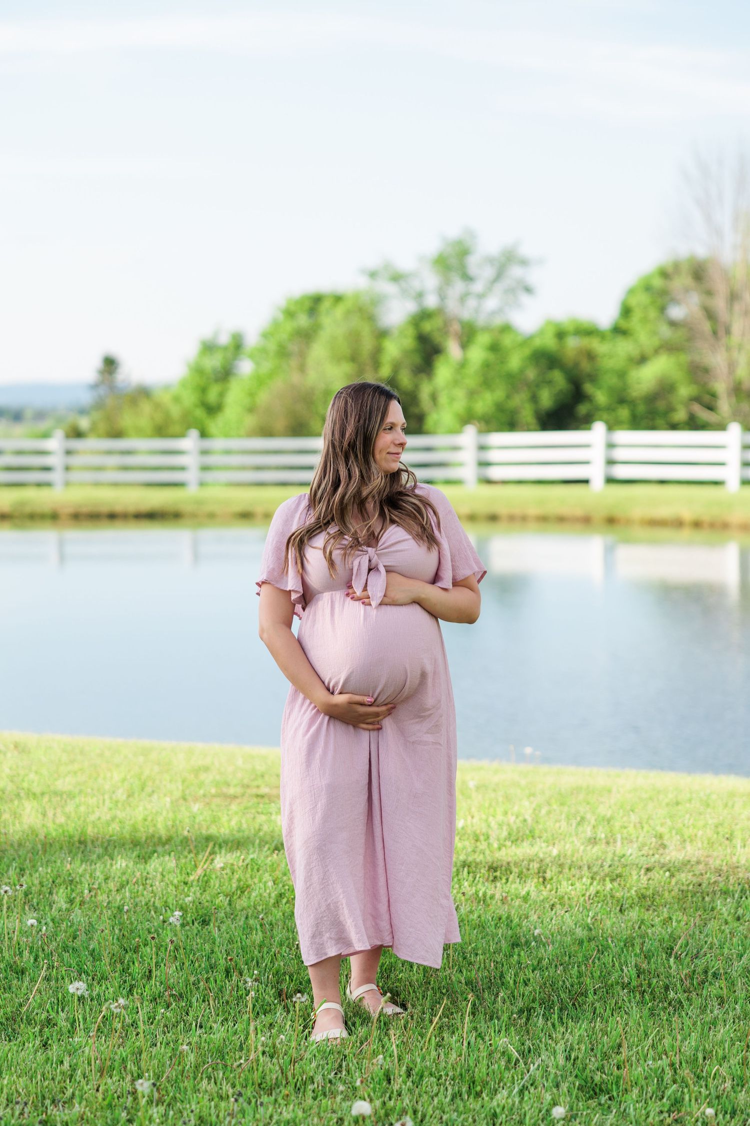Pregnant woman in pink dress gazing sideways by a serene pond outdoors.