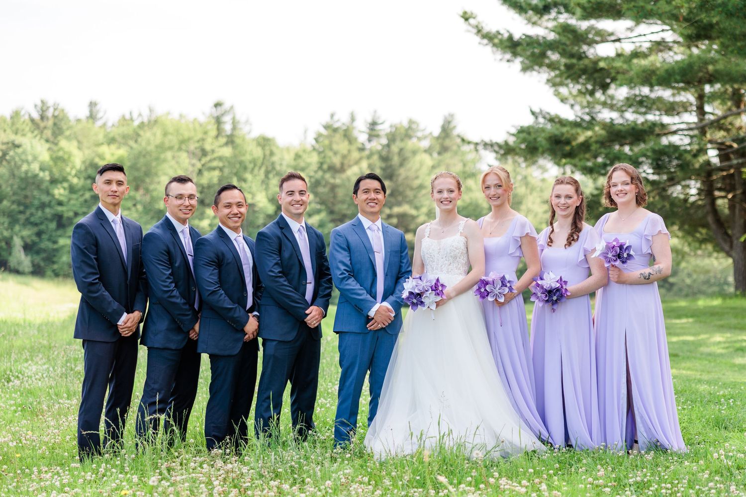 Wedding party poses in garden, groomsmen in navy suits, bridesmaids in lavender gowns.