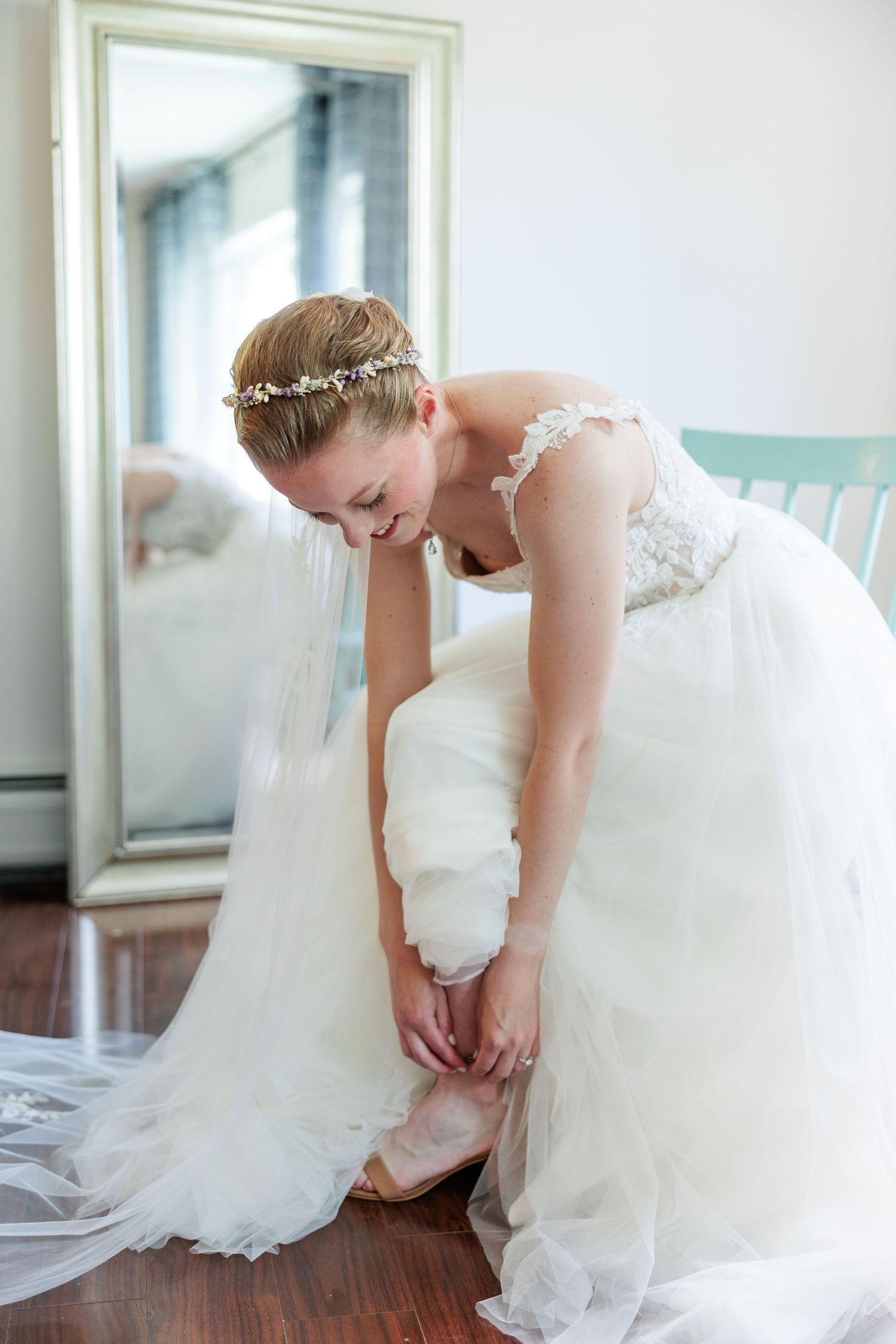 Bride in a lace tulle wedding gown with floral crown fastening her shoes beside a full-length mirror.