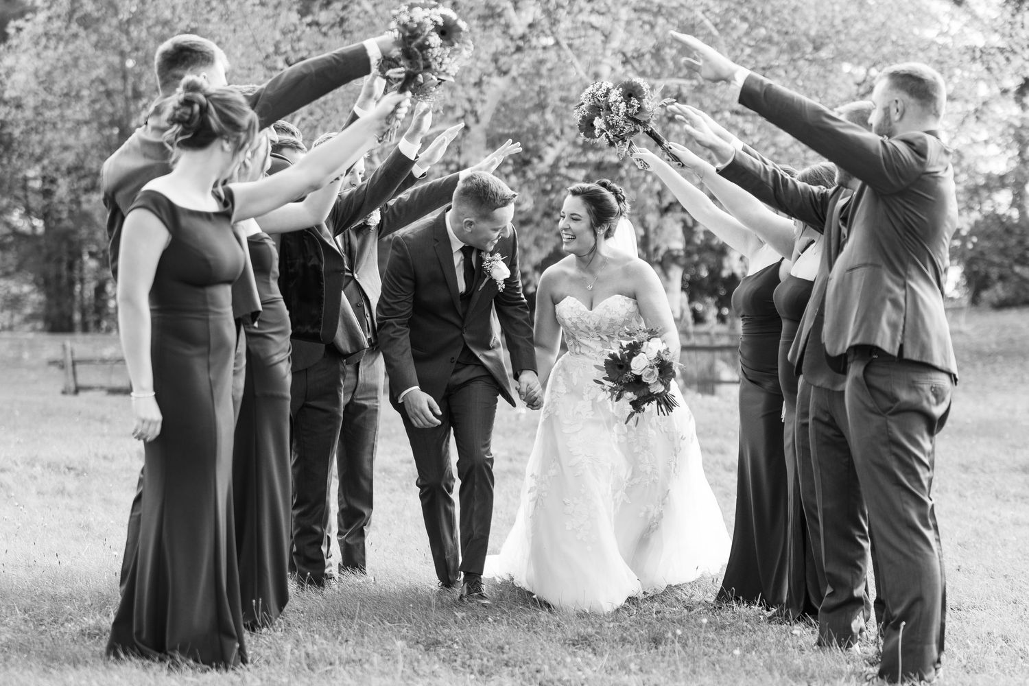 Black and white photo of bride and groom walking under arch of raised bouquets held by wedding party.