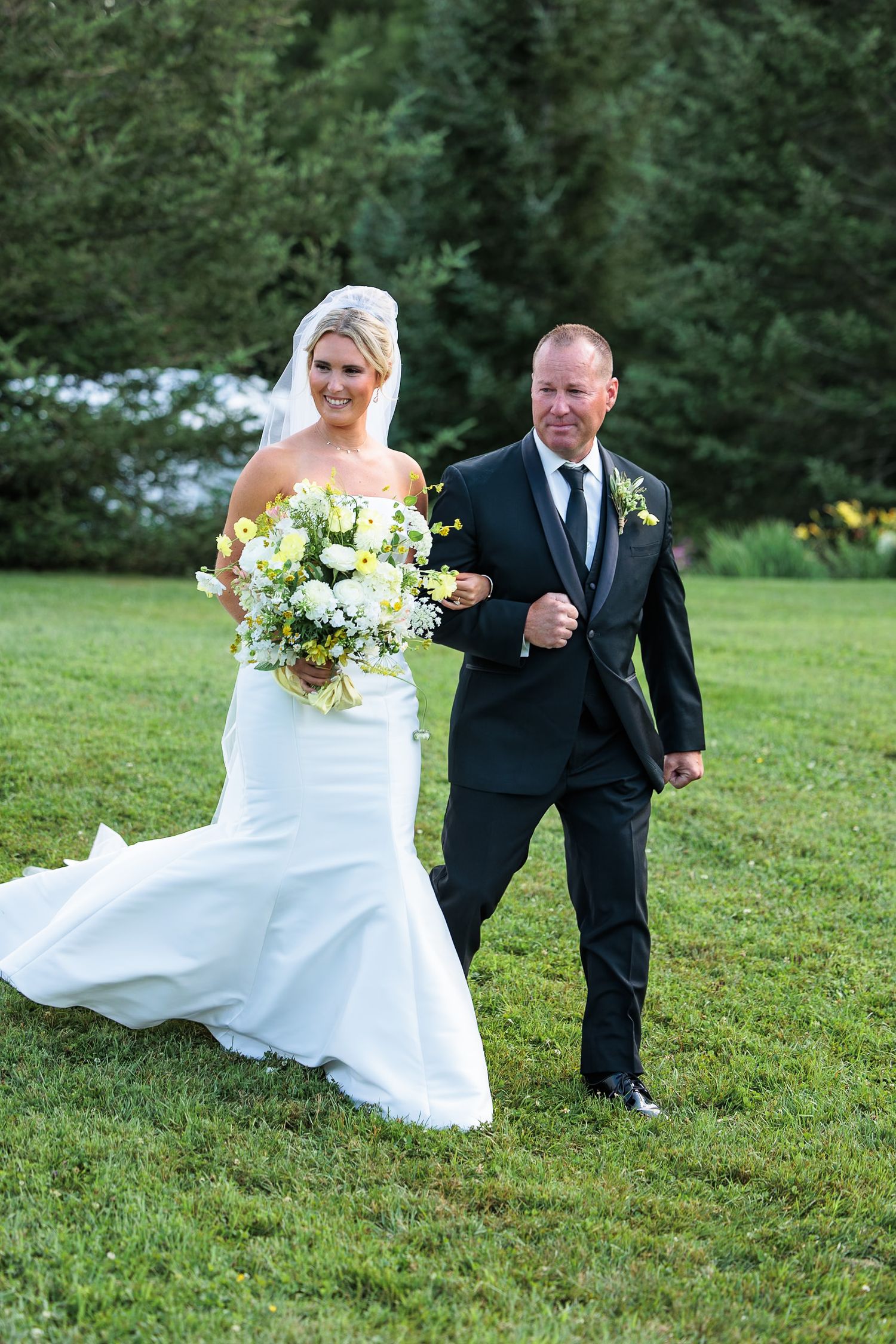 Bride in white mermaid gown walking with escort carrying yellow and white flower bouquet.
