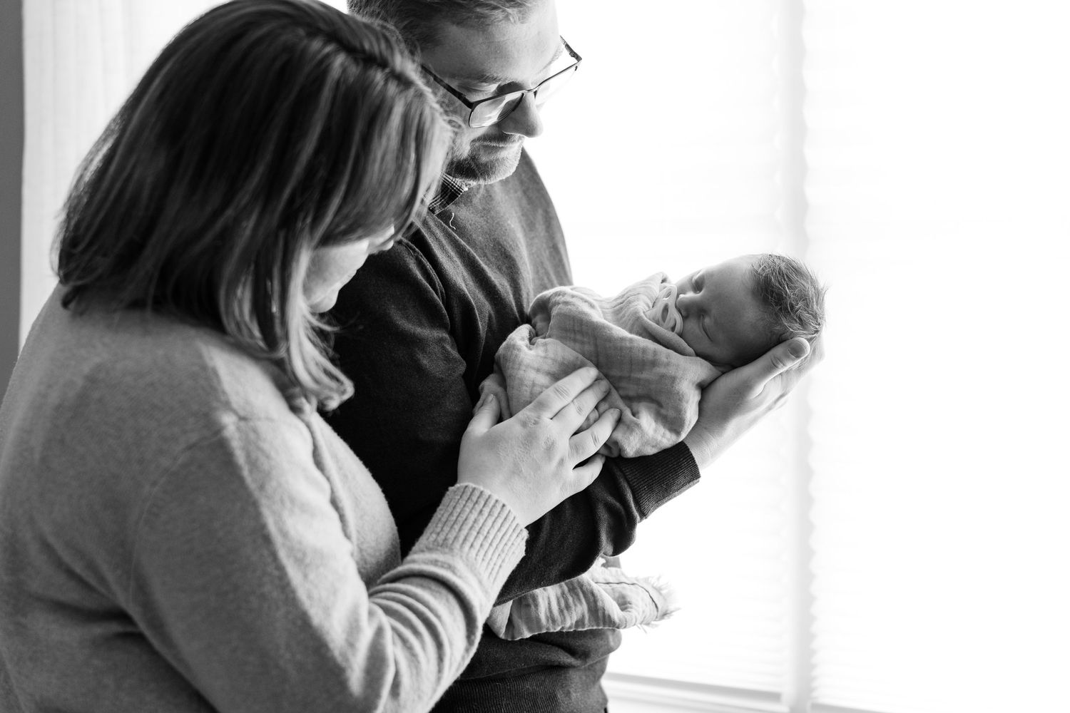 Black and white photo of new parents lovingly gazing at their newborn baby by a bright window.
