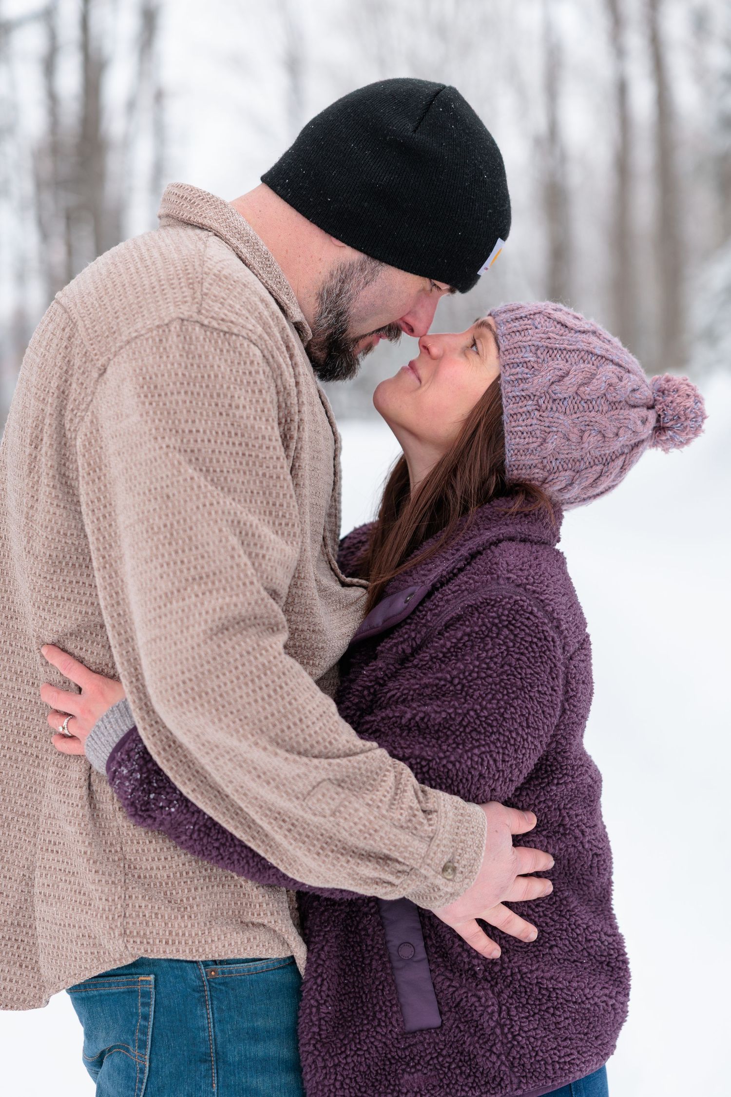 Couple shares a romantic kiss outdoors in winter, wearing cozy sweaters and knit hats in a snowy setting.