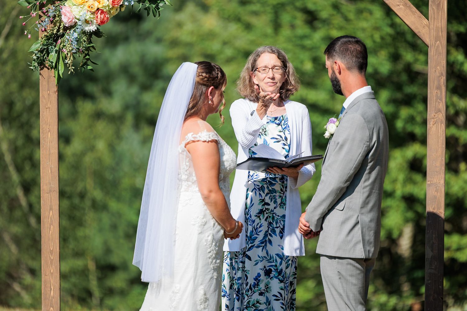 Officiant reads vows as bride and groom hold hands under floral-decorated wooden arch at outdoor ceremony.