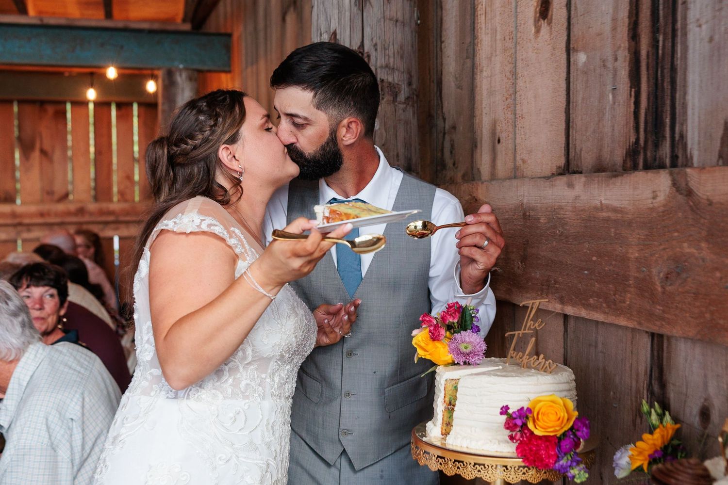 Bride and groom sharing a playful cake tasting moment inside a rustic wooden barn reception.