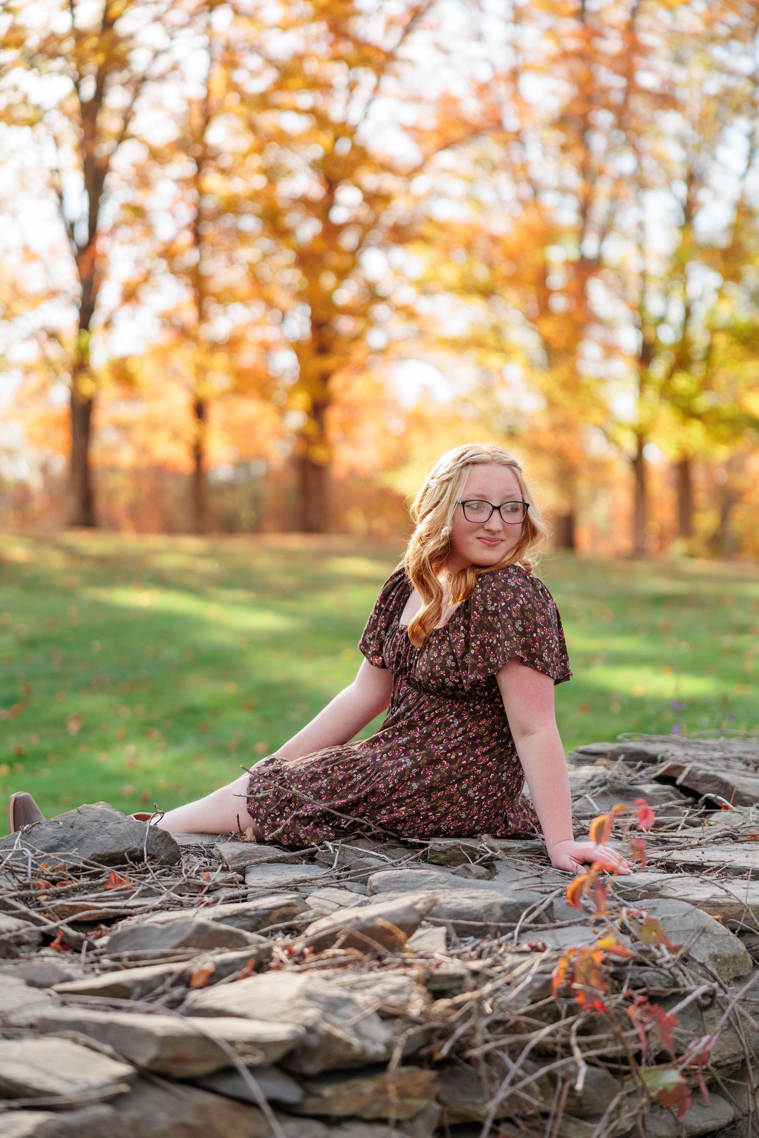 Young blonde woman in floral dress sitting on stone wall surrounded by colorful autumn foliage in a park.