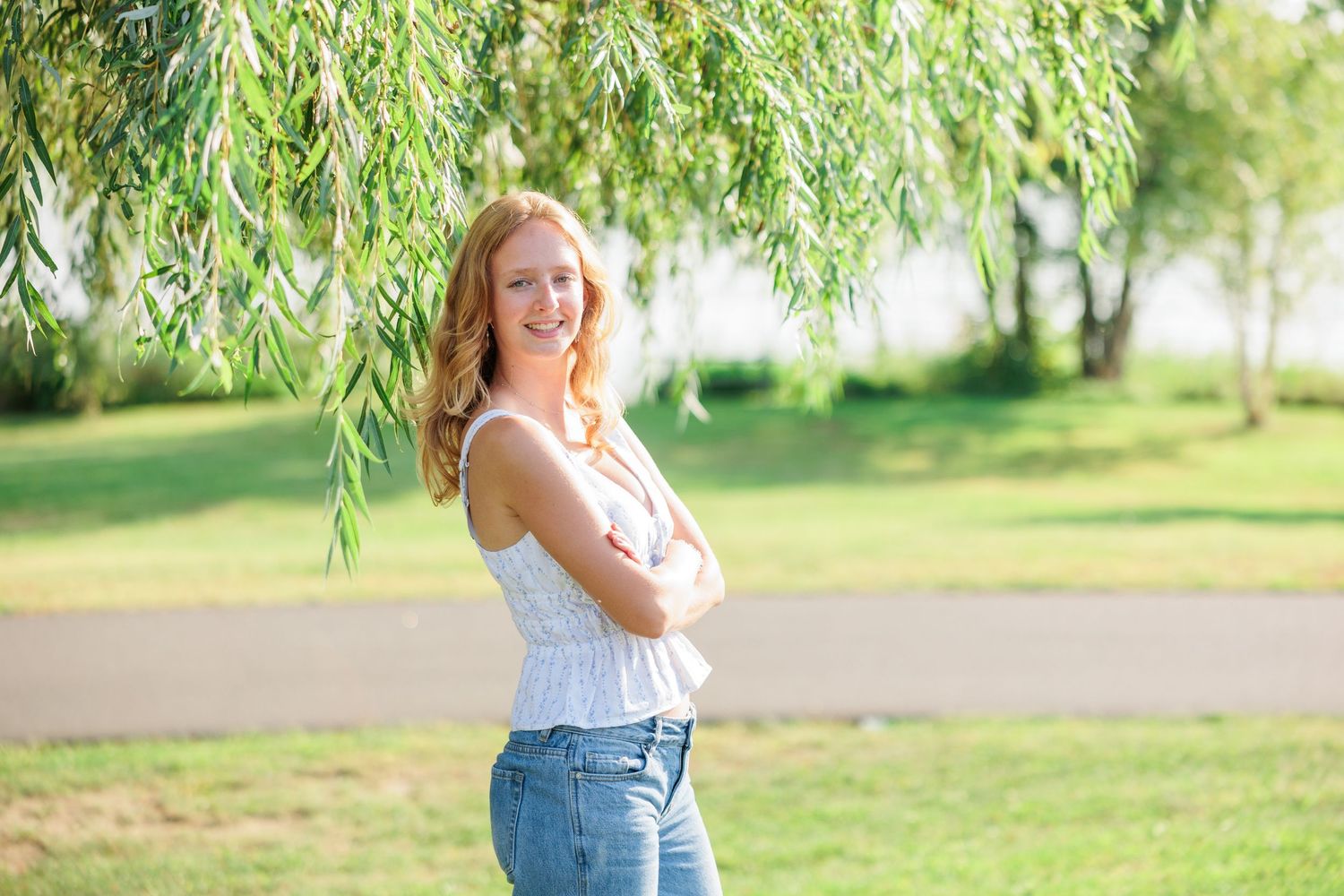 Young woman with red hair smiling outdoors, arms crossed, wearing white top and jeans under a willow tree.