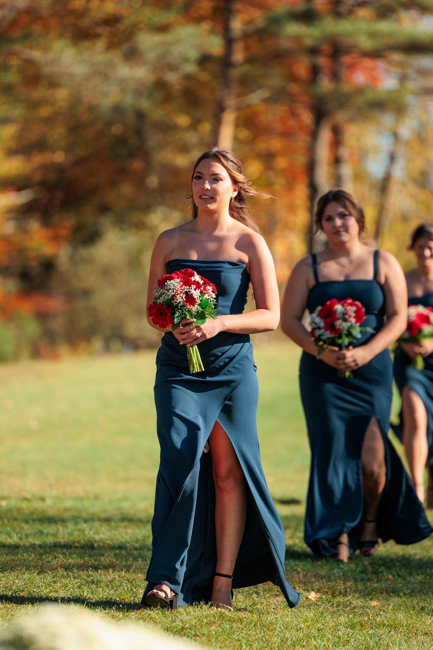 Bridesmaids in teal dresses holding red bouquets stand in a line on grass during an autumn wedding ceremony.